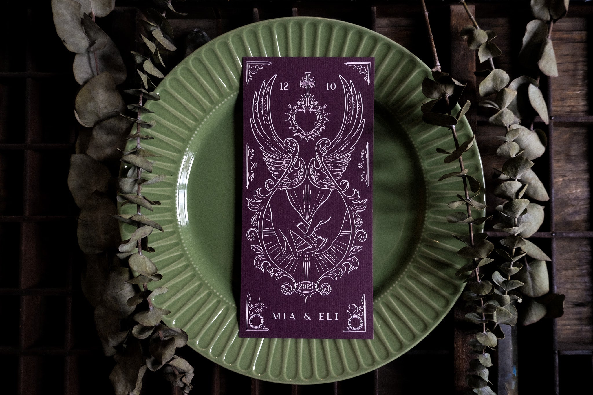 Luxury wedding menu card with ornate white tarot-inspired design on rich card, styled atop a green plate with dried eucalyptus; mystical, romantic UK stationery.