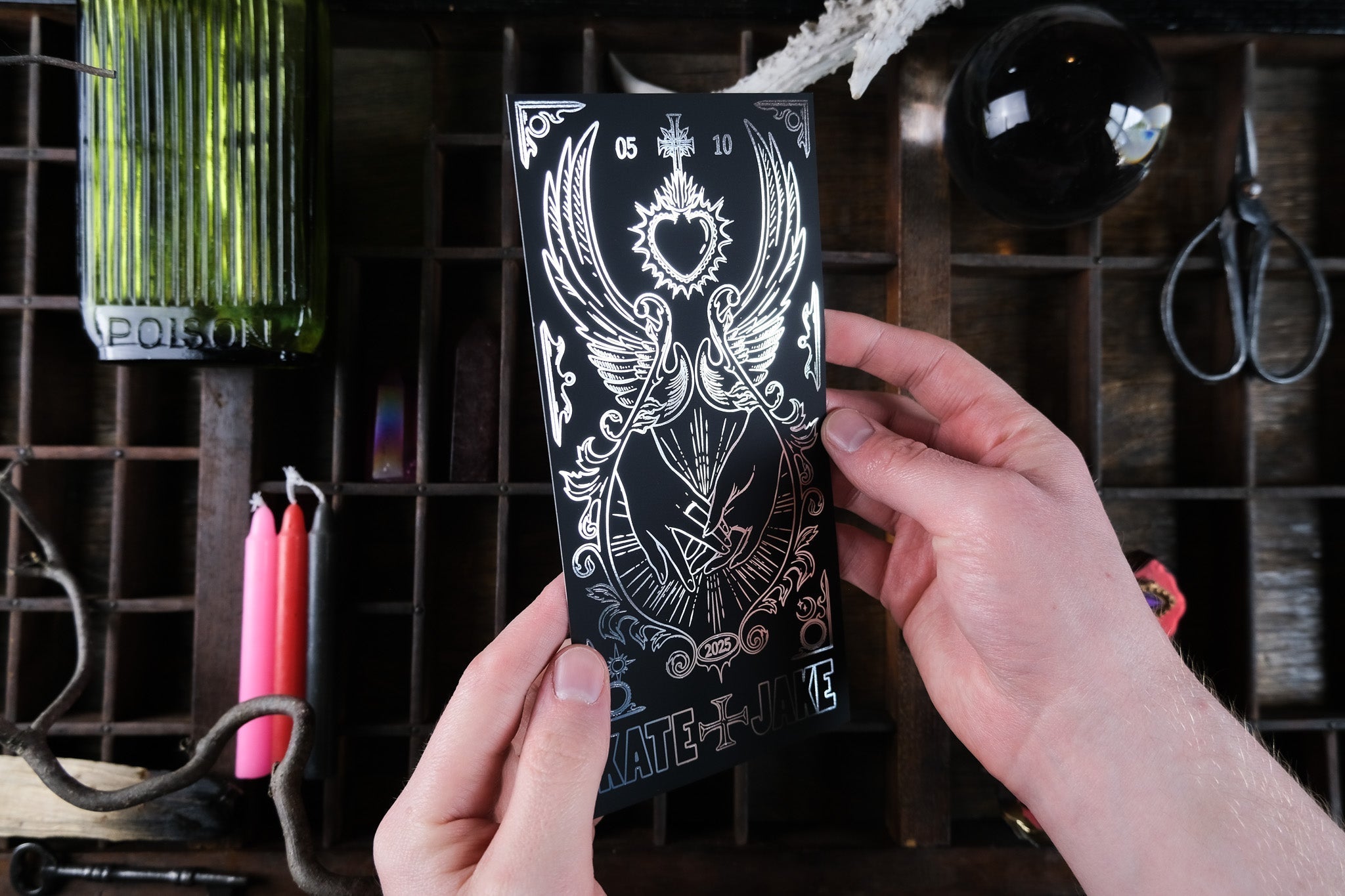 A hand displays a luxury foiled gothic save the date with celestial symbols, amid red candles, green apothecary bottle, and crystal ball.