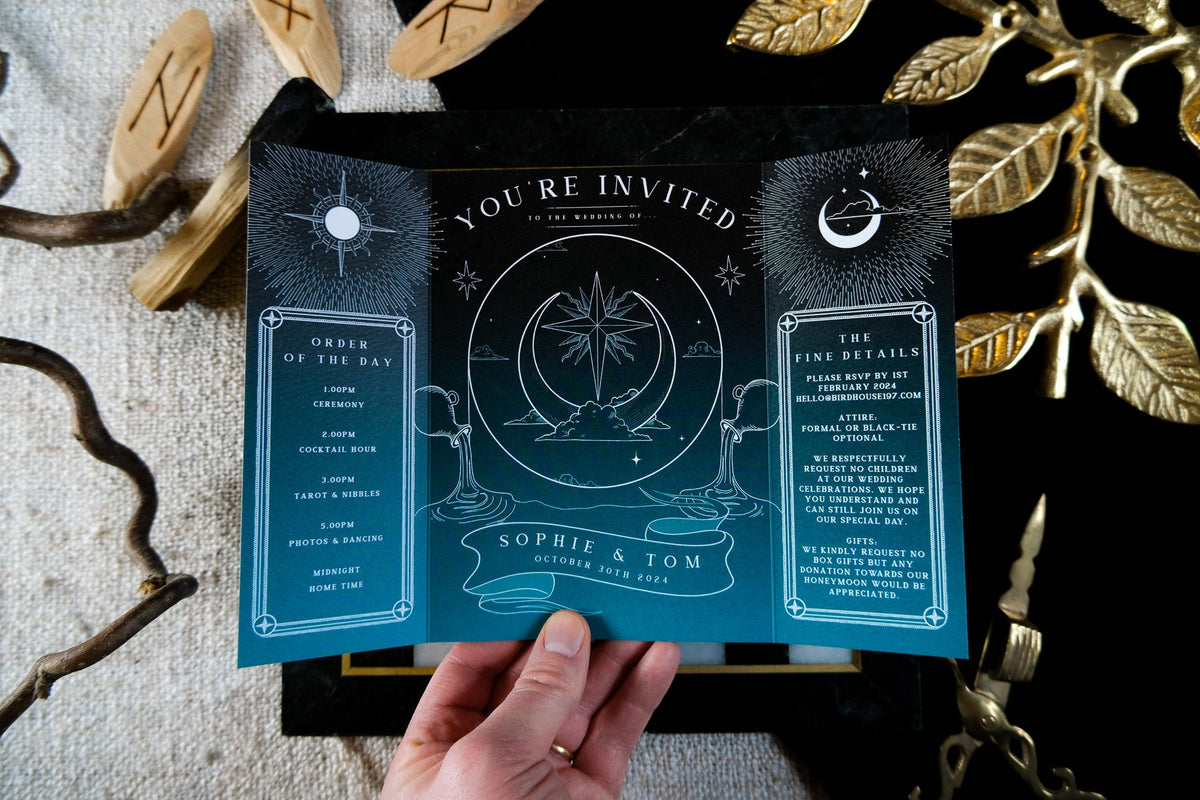 A hand holds a luxury celestial gatefold wedding invite in deep blue, adorned with stars, constellations, and mystical moon motifs.