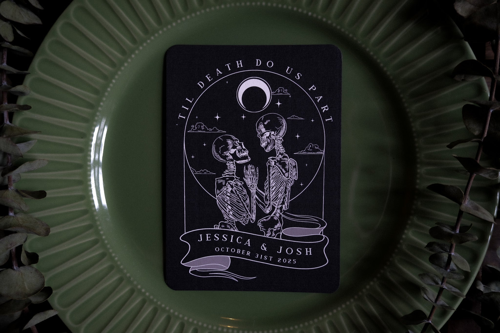 Luxury gothic wedding menu card with a skeleton couple beneath a crescent moon, set on green china amid lush leaves; UK stationery.