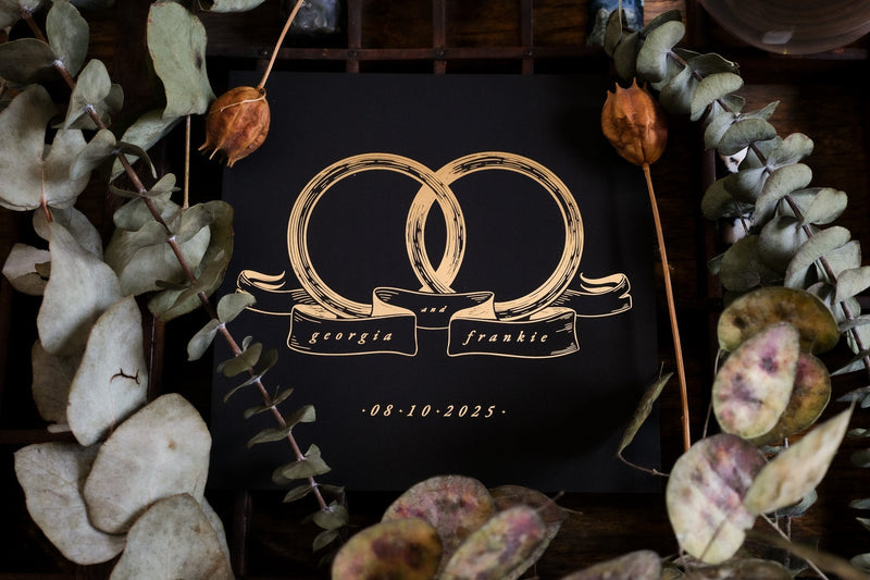 Luxury foiled save the date card featuring interlocking gold rings on black, with dried florals; dramatic, romantic UK wedding stationery.