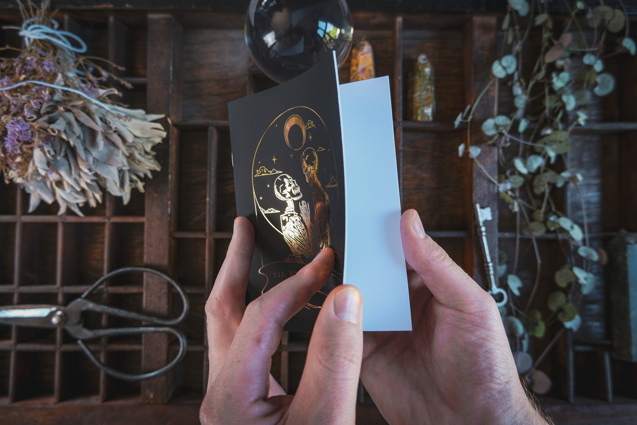 A person opens a luxury black vow book with gold foil design, styled in a gothic and dramatic look on a wooden table with vintage decor.