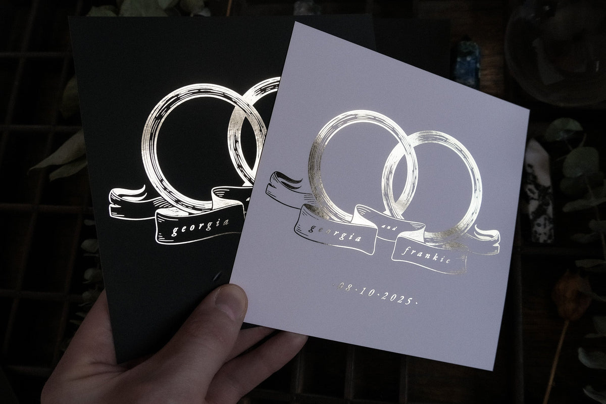 A hand displays two luxury gold foil wedding save the date cards with intertwined rings, a ribbon accent, and elegant script detail.
