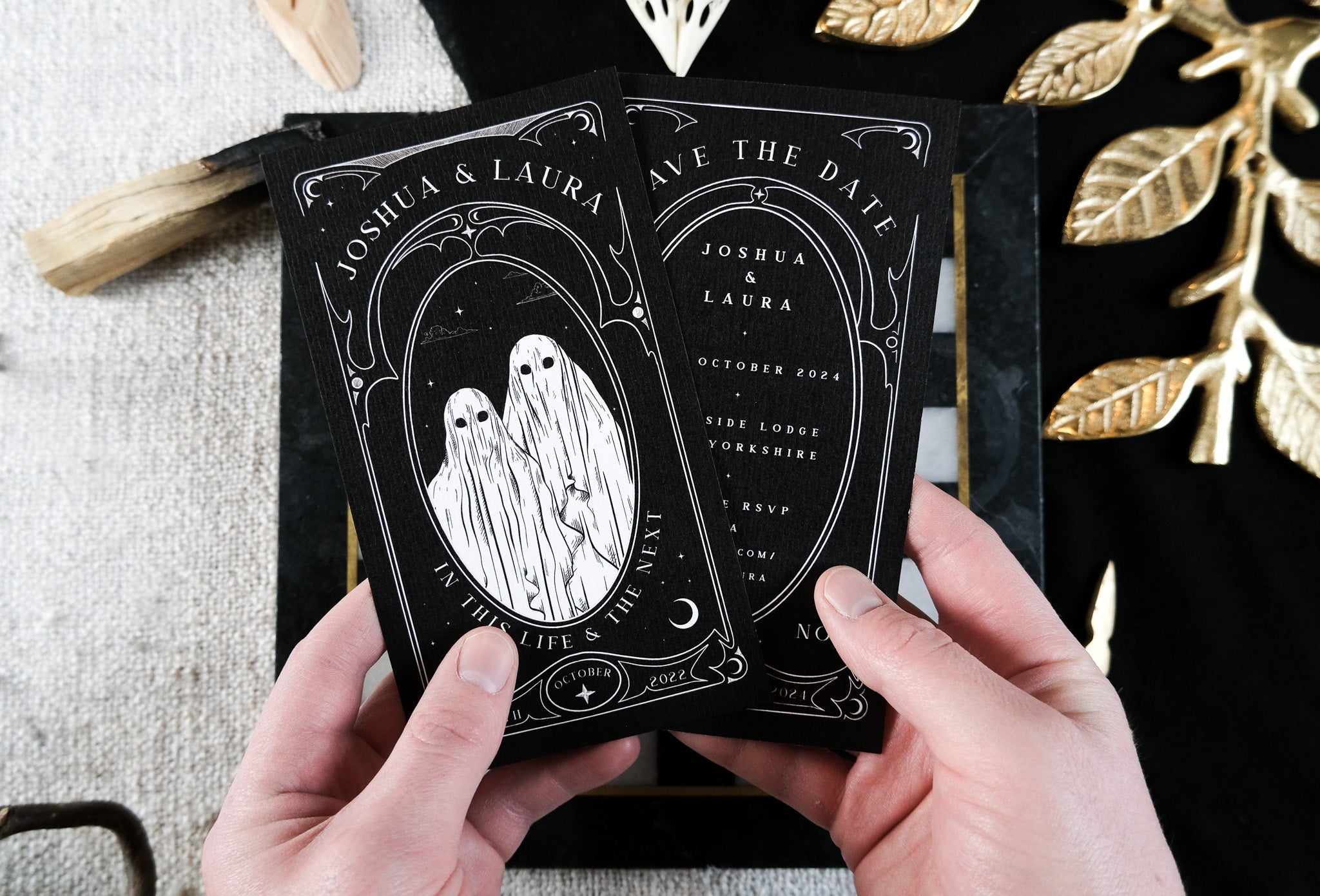 Two hands hold a luxury UK gothic wedding invitation featuring ornate white design, ghost motifs, and dramatic, mystical details.