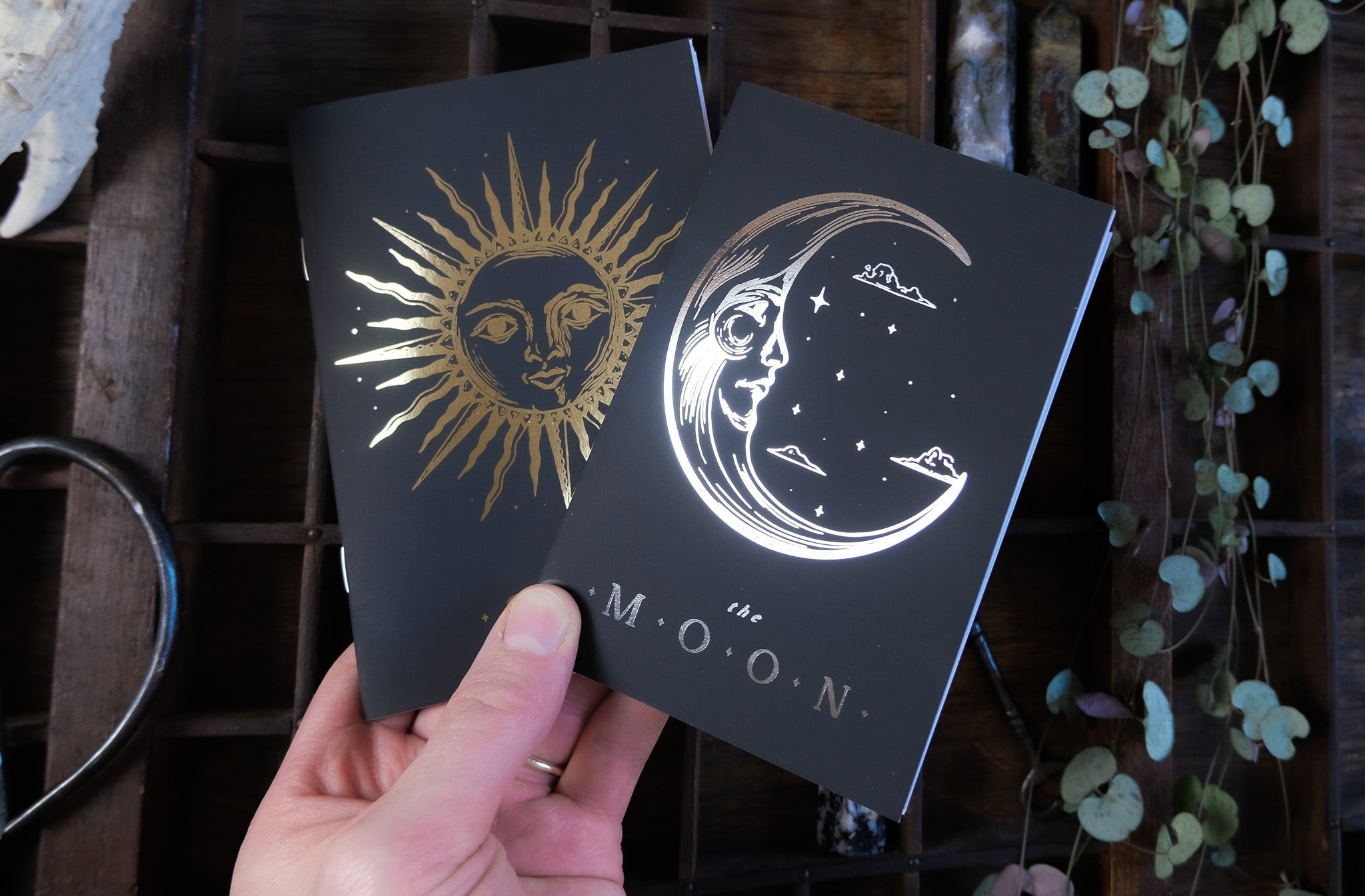 A hand displays luxury gold and silver foiled celestial vow books with gothic sun and moon designs, set amid plants and vintage wood.