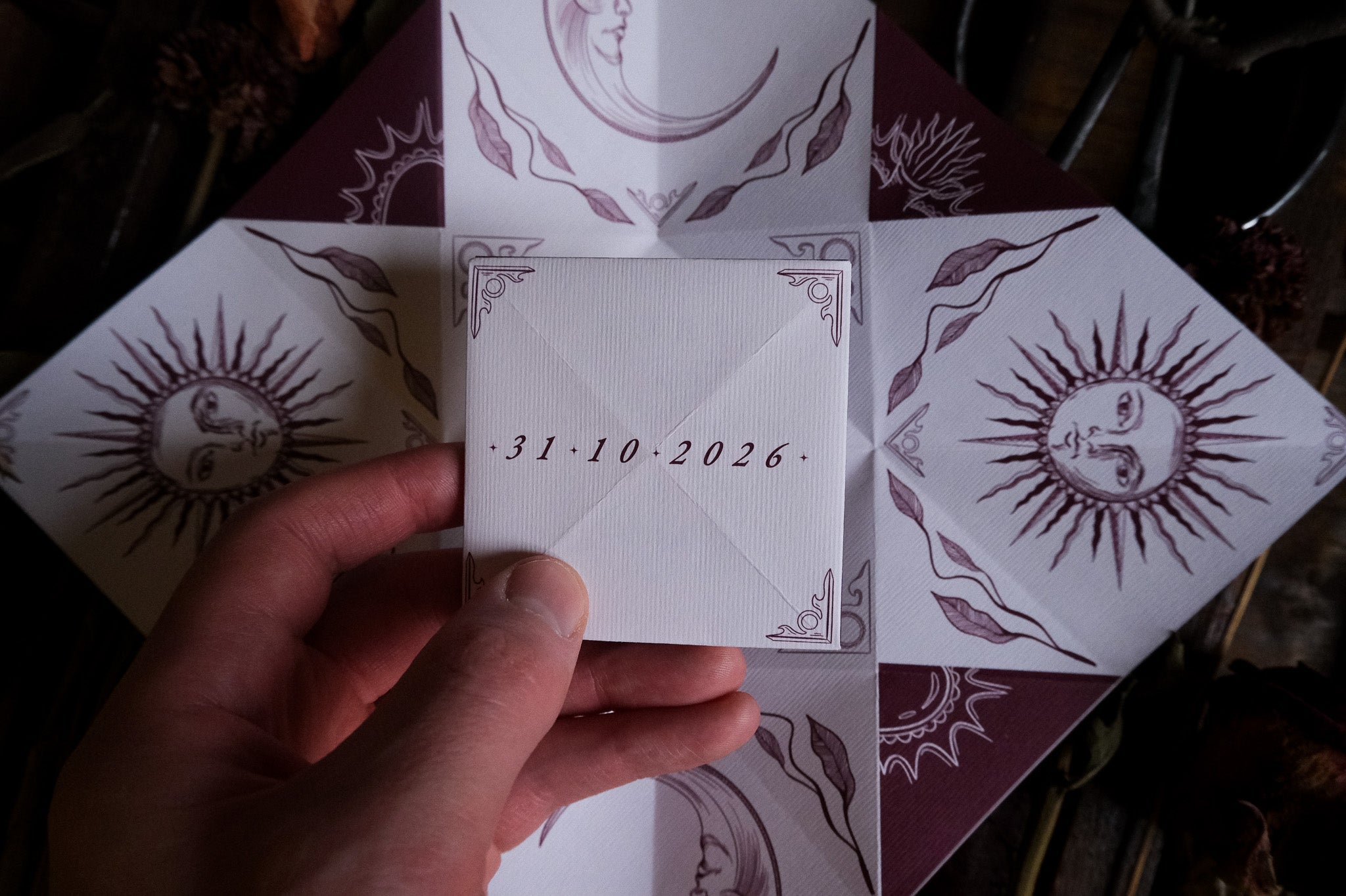 A hand holds a luxury celestial wedding card with vintage sun and moon motifs in deep purple, styled amid dried florals and rustic boho touches.