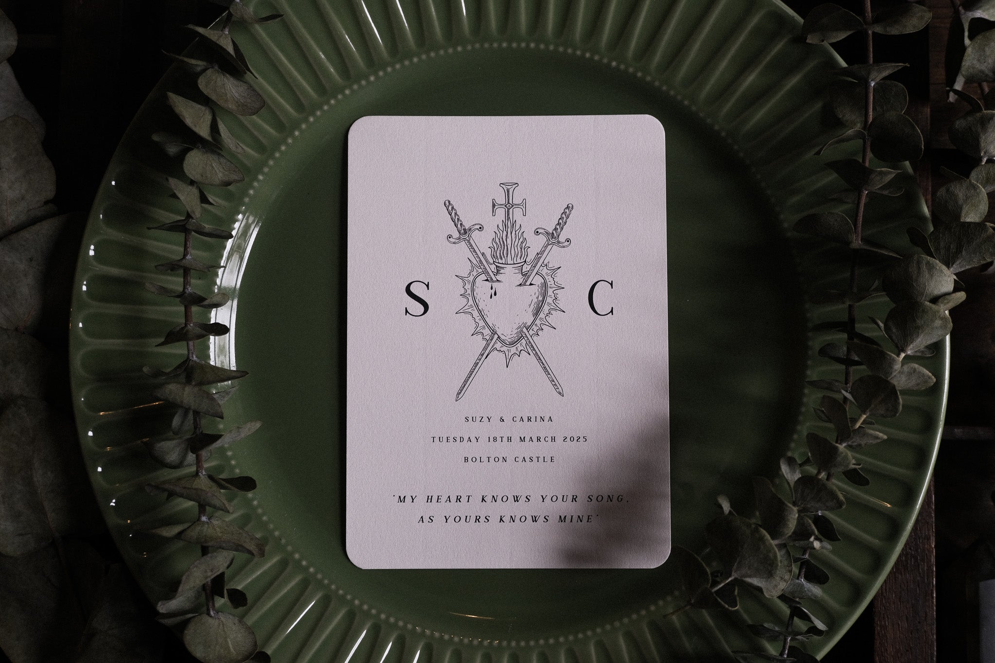 Luxury gothic wedding menu with bleeding heart and crossed swords artwork on textured card, styled with eucalyptus on vintage green plate.