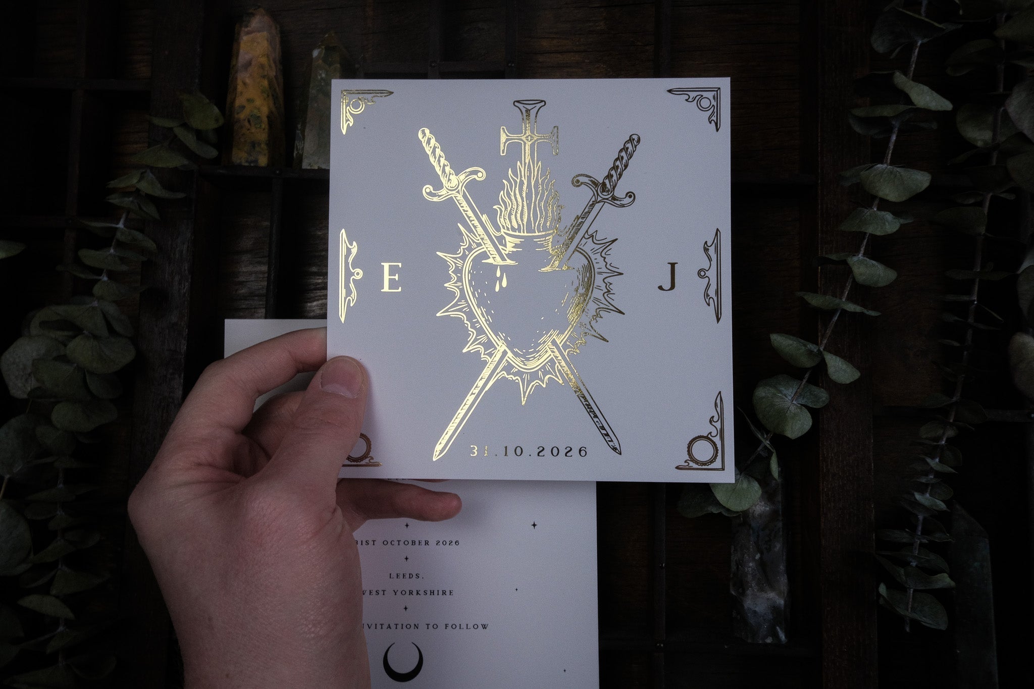A hand displays a square, gothic foiled save the date card with gold dramatic motifs; luxury UK wedding stationery amid plants and bottles.