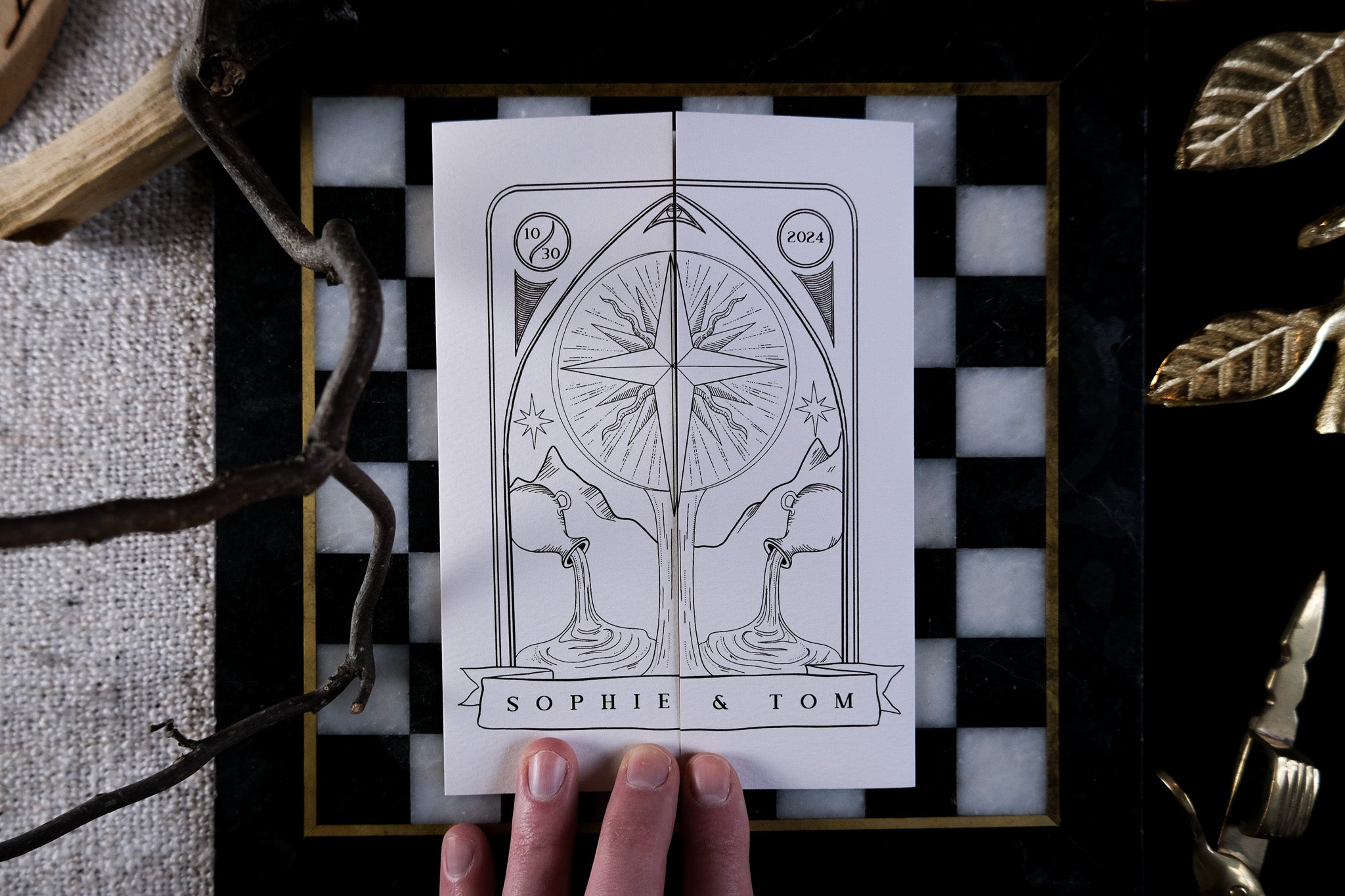 A hand holds a luxury celestial gothic wedding gatefold invitation with ornate stars, mirrored figures, foil details, on a chessboard.