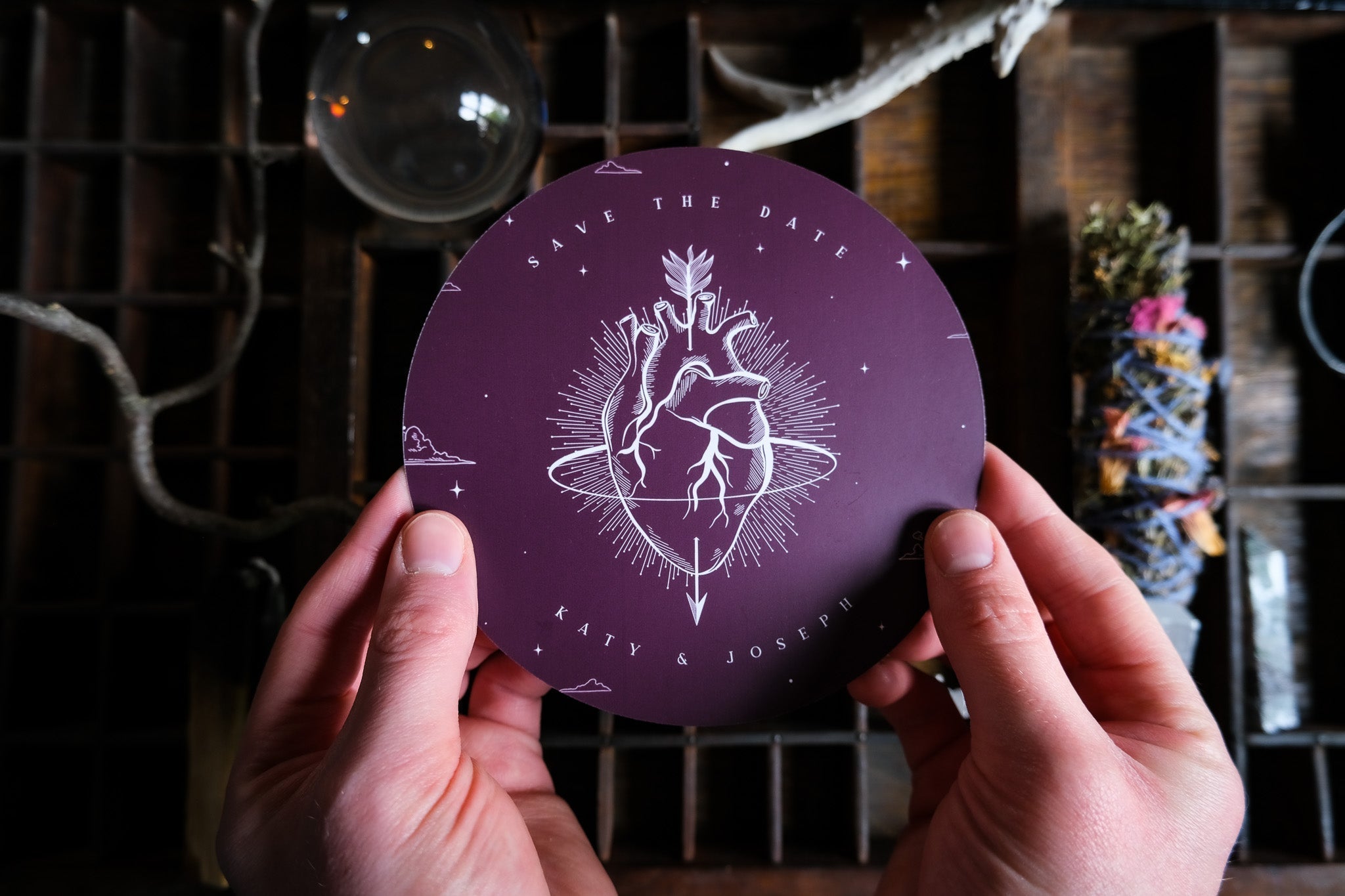 A hand holds a luxury gothic circular save the date card, featuring a white anatomical heart on deep purple with geometric, dramatic accents.