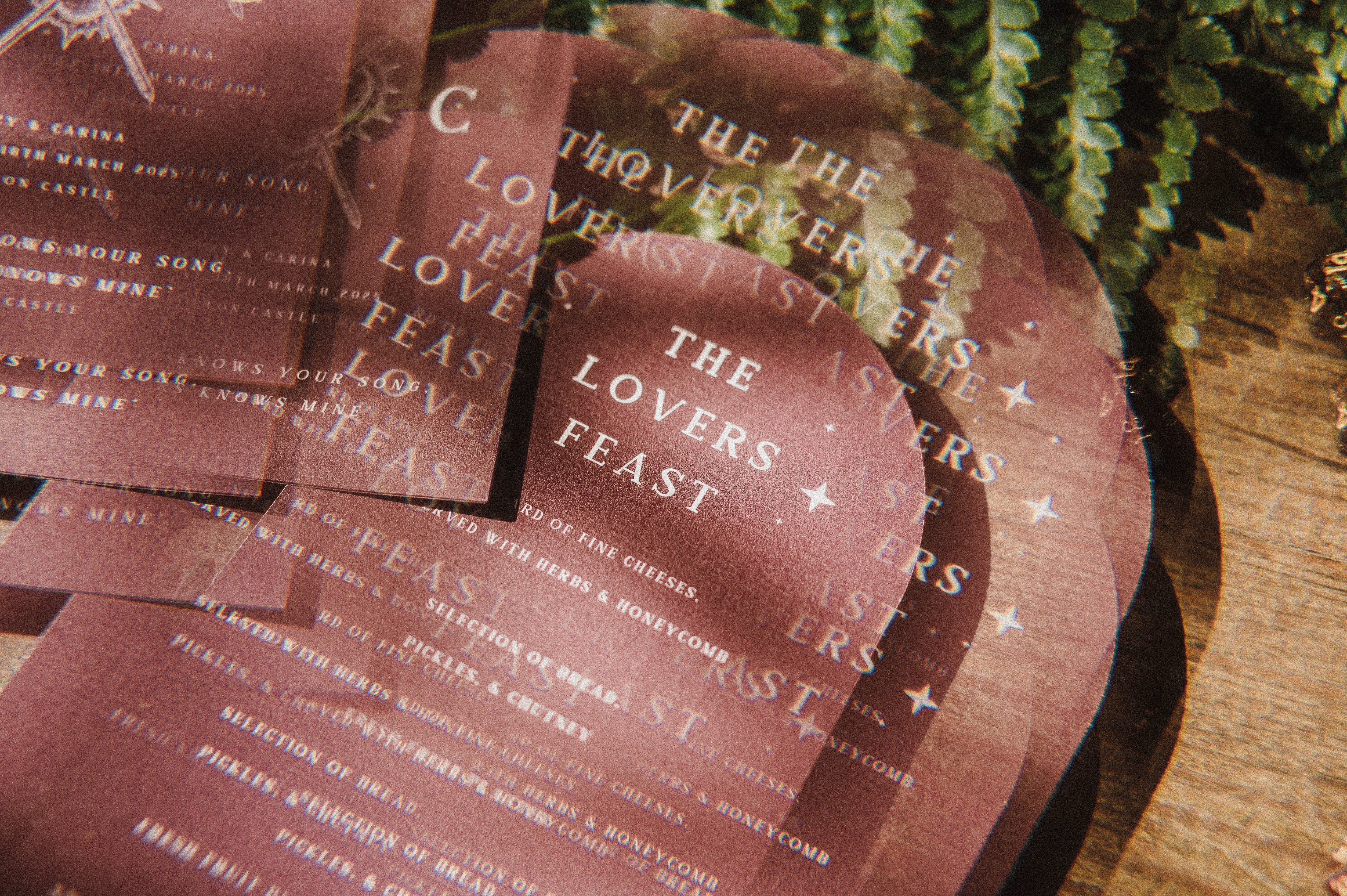 A close-up of luxury maroon arched wedding menus with gold foil celestial motifs, layered on wood beside fern leaves; romantic UK stationery.