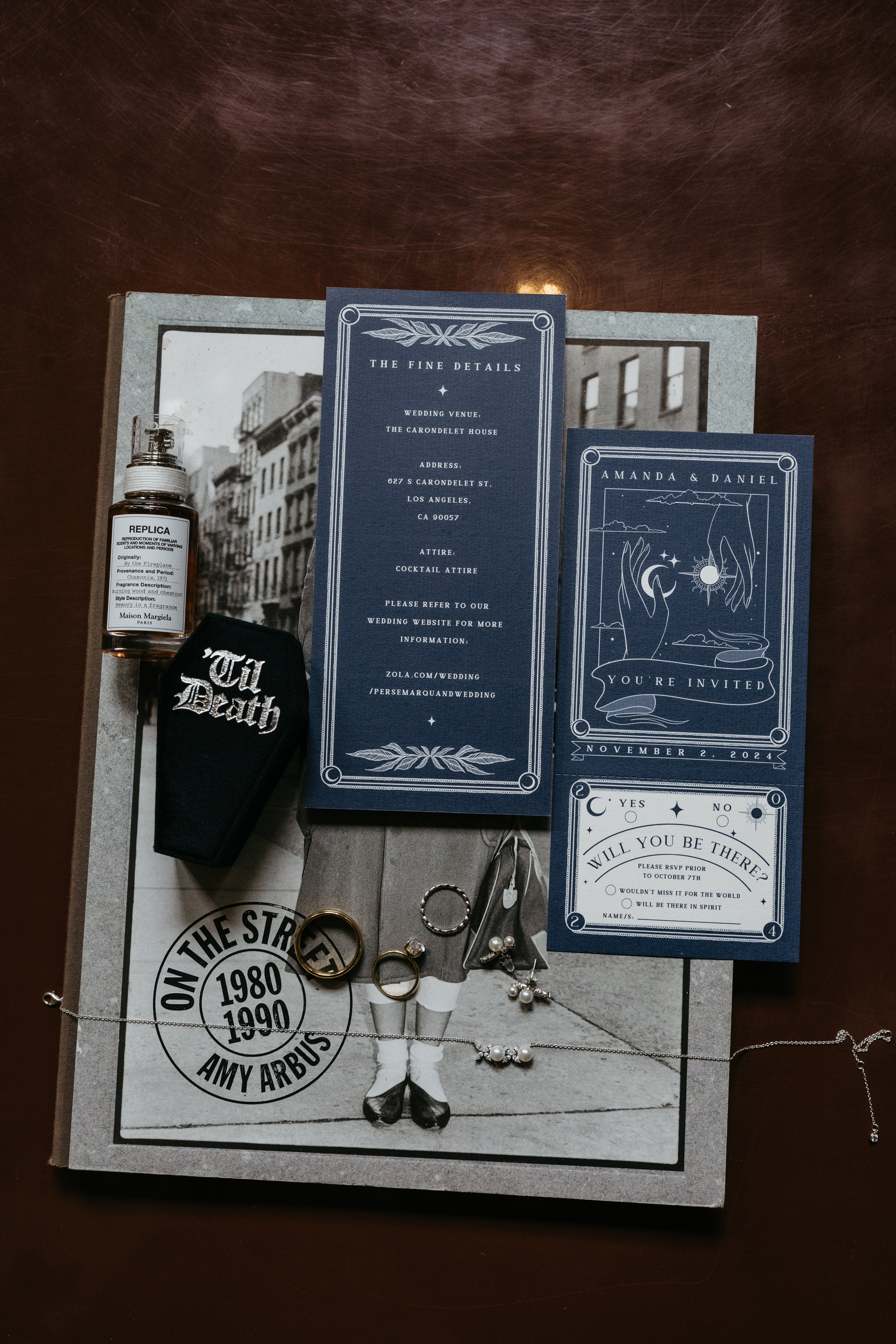 Flat lay of luxury wedding stationery with a dramatic gothic design, vintage photo book, perfume bottle, and eclectic details on brown.