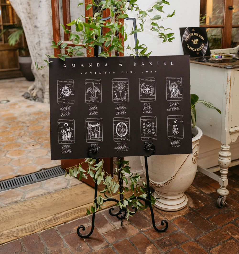 Black tarot card seating chart on an easel with greenery in an indoor wedding venue
