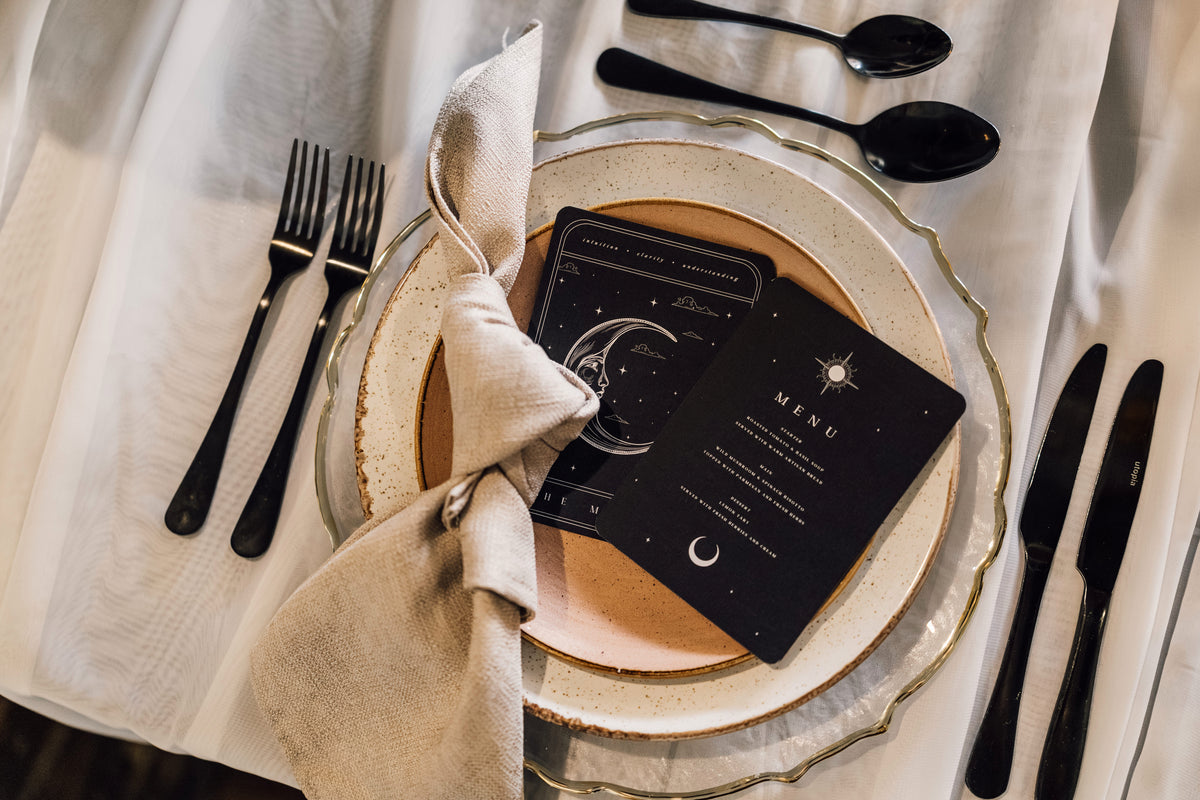 Celestial-style black menu and table card with white illustrations set on a white tablecloth alongside a layered place setting.