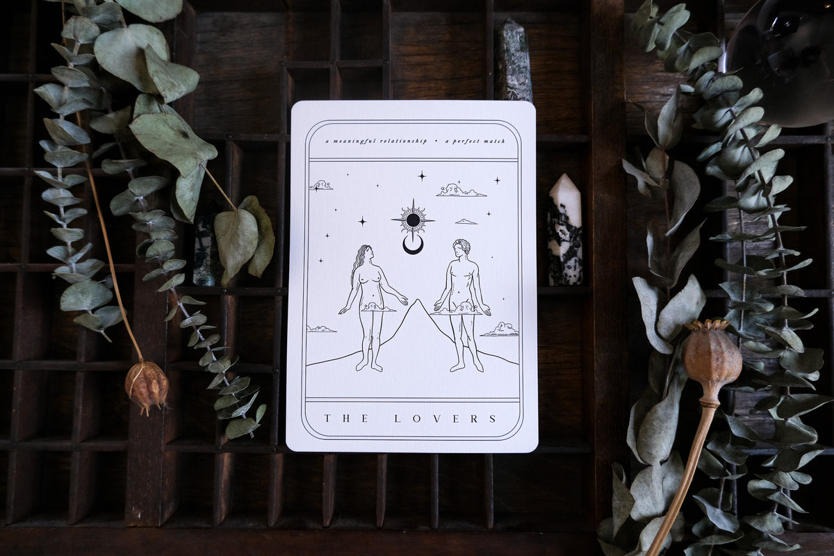 A luxury tarot-inspired wedding stationery card with a minimalist celestial design, set on wood amid dried leaves and crystals.