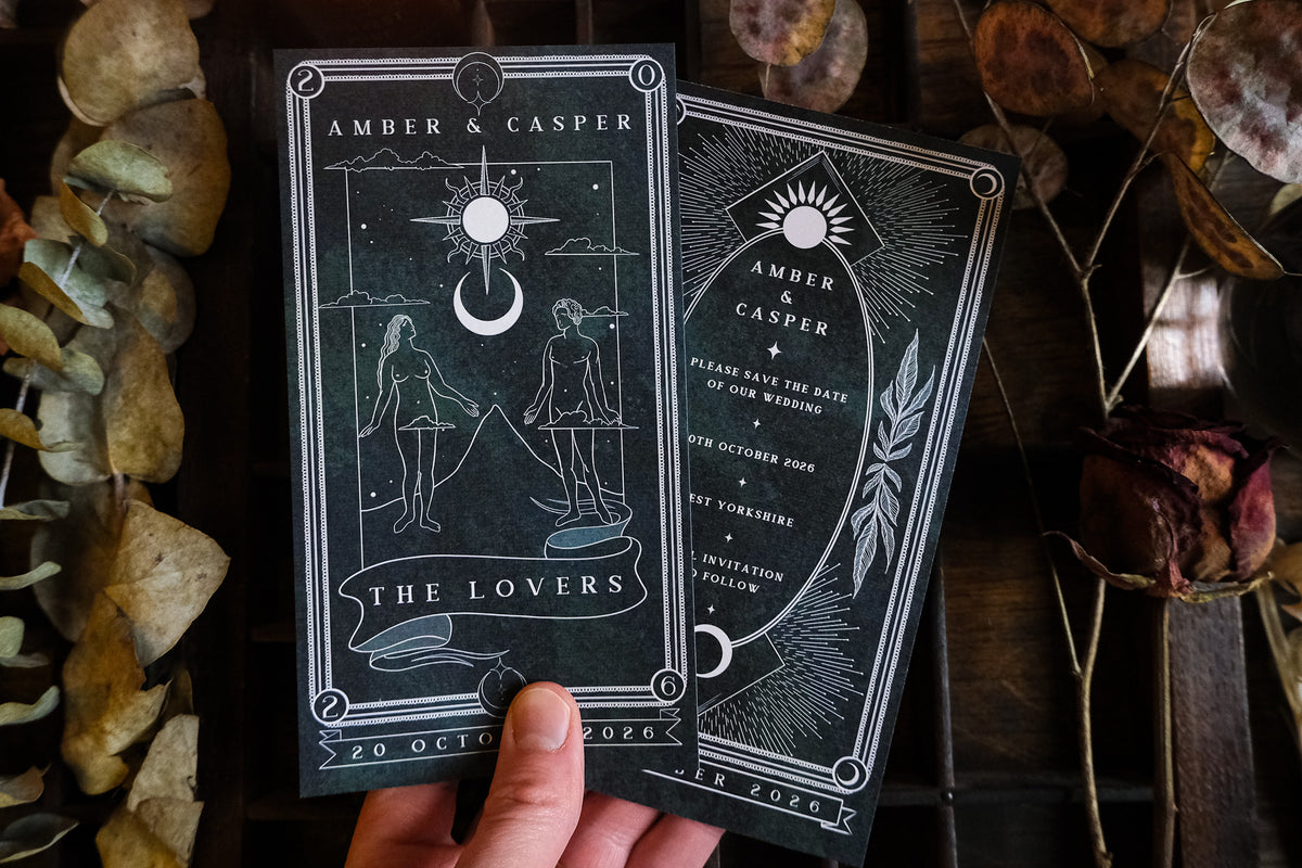 A hand displays two luxury wedding stationery pieces from a UK collection, designed in a gothic and mystical style with intricate tarot-inspired illustrations and subtle celestial motifs. The save the date cards feature dramatic, alternative artwork printed on premium stock. In the background, dried leaves enhance the romantic, vintage atmosphere, reflecting an enchanting boho mood perfect for personalised celebrations.