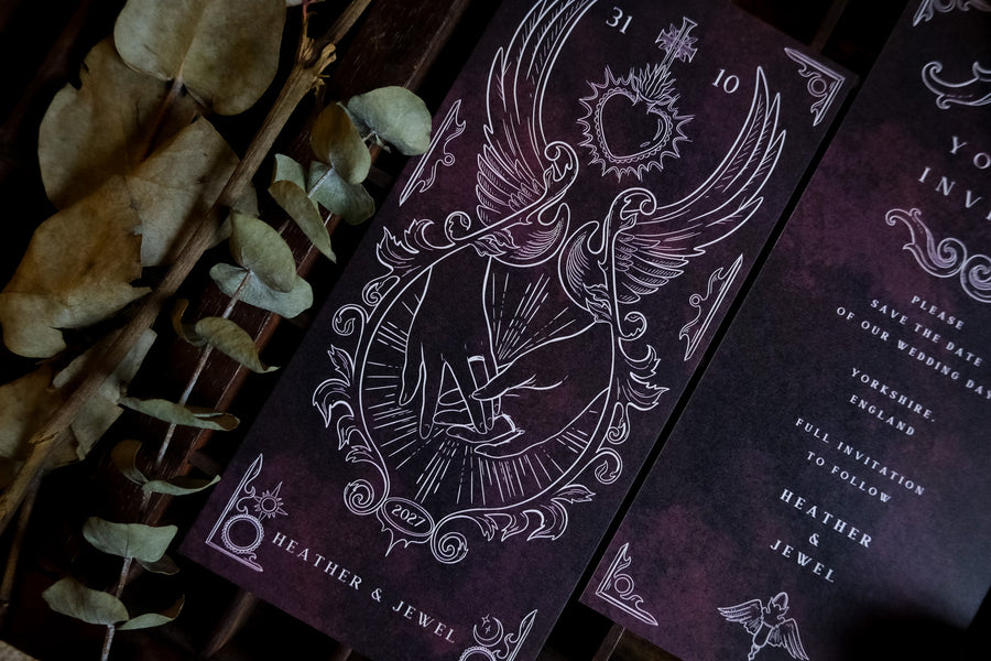 Purple and black tarot-inspired save the date for Heather & Jewel, featuring a winged hand and sacred heart illustration with a 2027 wedding date detail, styled with dried eucalyptus.
