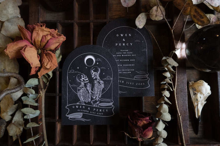 Arched black save the date for Gwen & Percy featuring a skeleton lovers illustration beneath a crescent moon, styled with dried roses and eucalyptus.