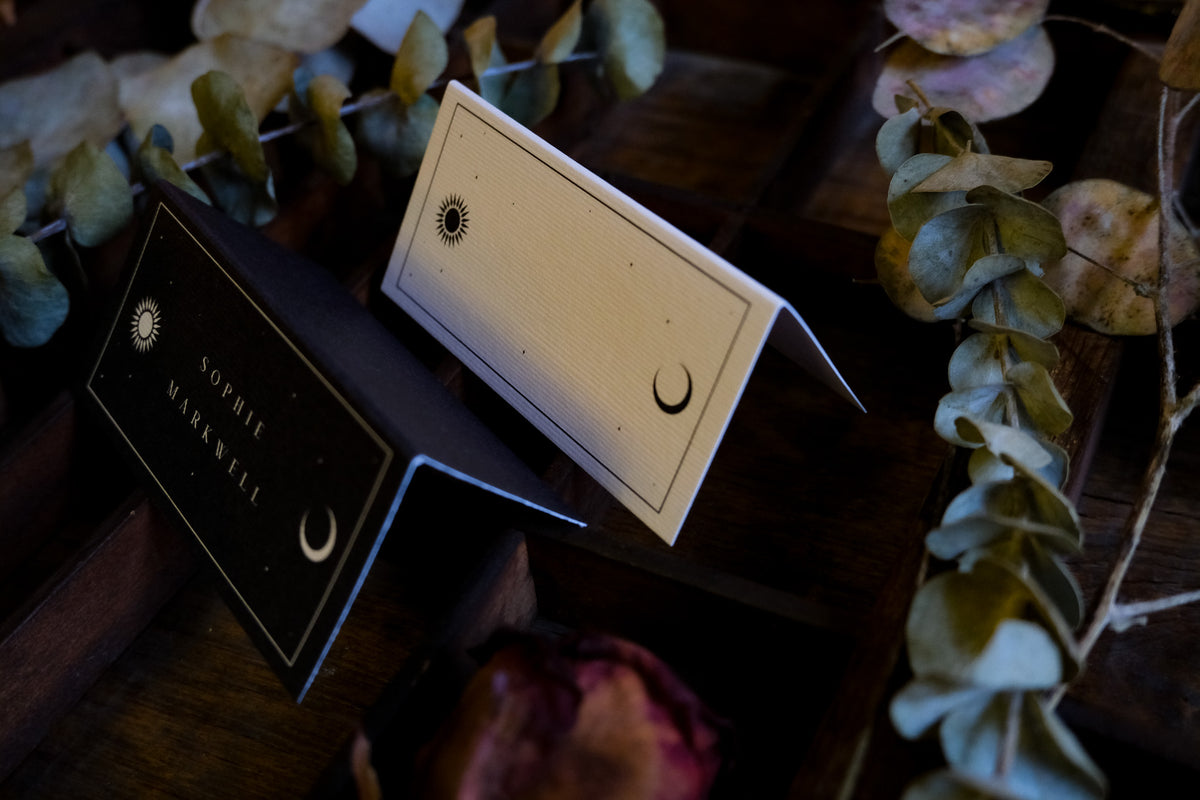 Luxury celestial wedding place cards, one black and one white, with mystical designs on wood and dried botanicals; gothic, alternative style.