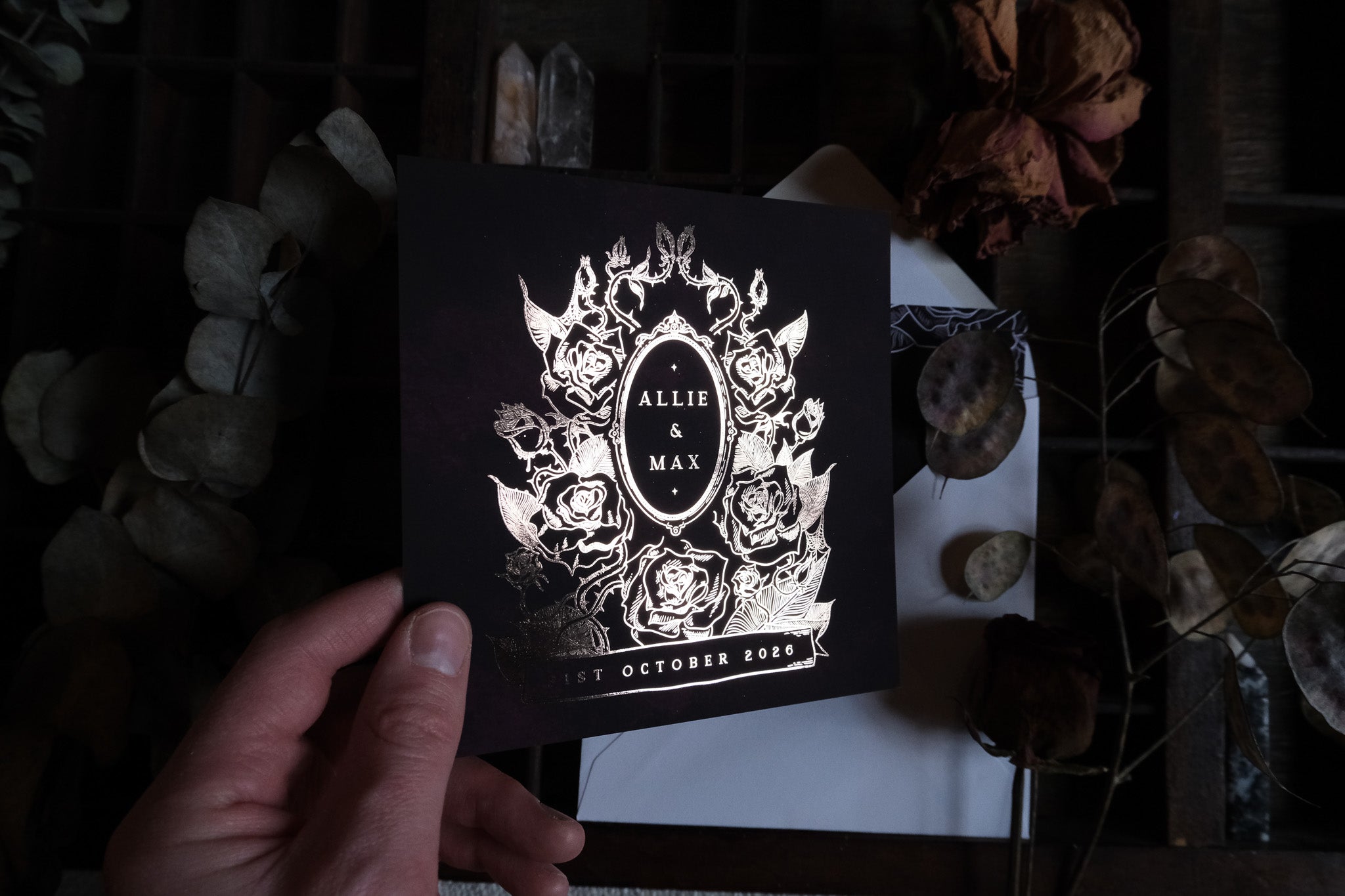 A hand displays a luxury black gothic wedding stationery card with silver foil florals and crown, beside dried flowers and envelope.