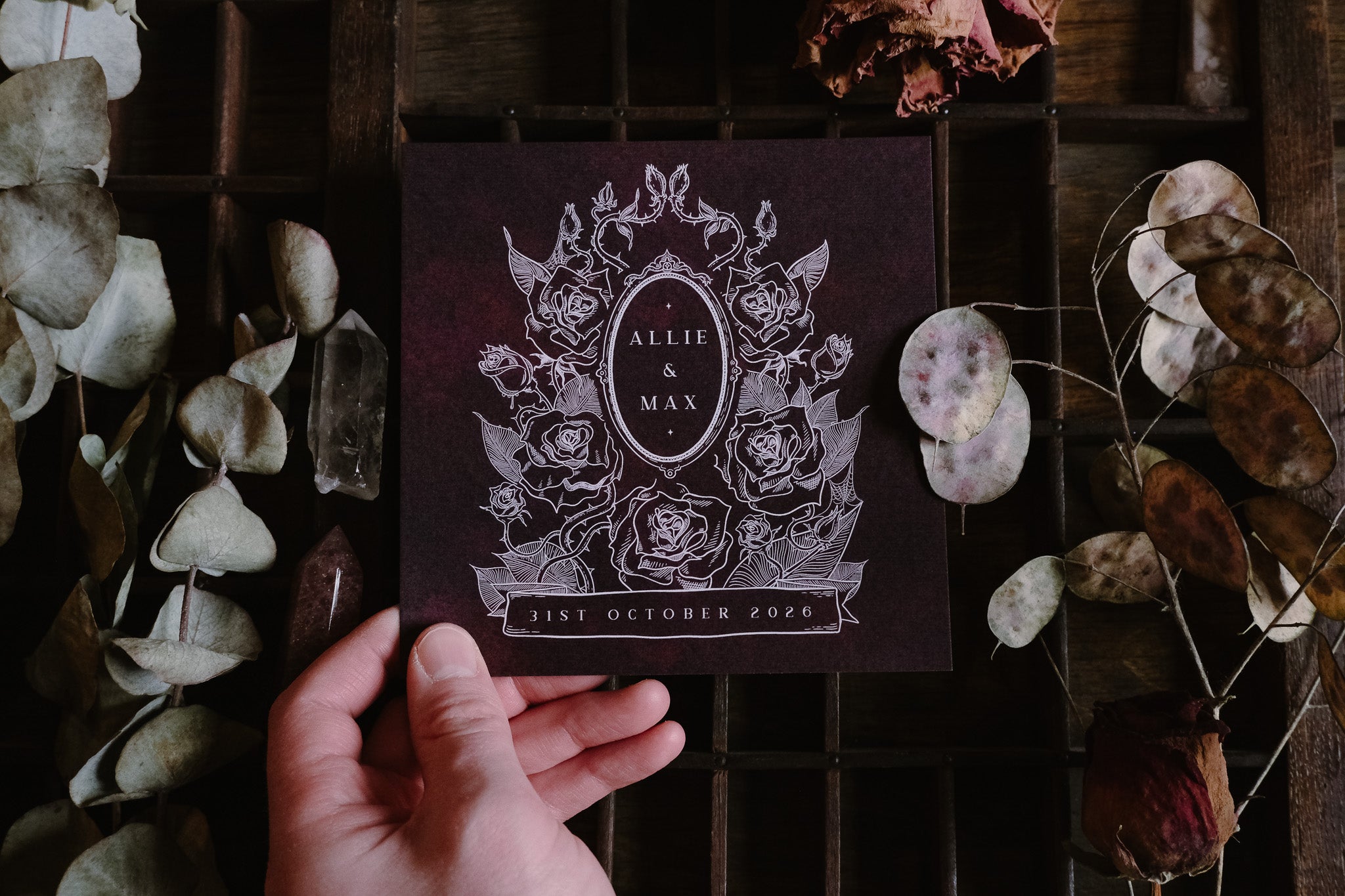 A hand holds a dramatic, gothic-inspired luxury save the date card with dark floral design, amid dried botanicals on rustic wood.