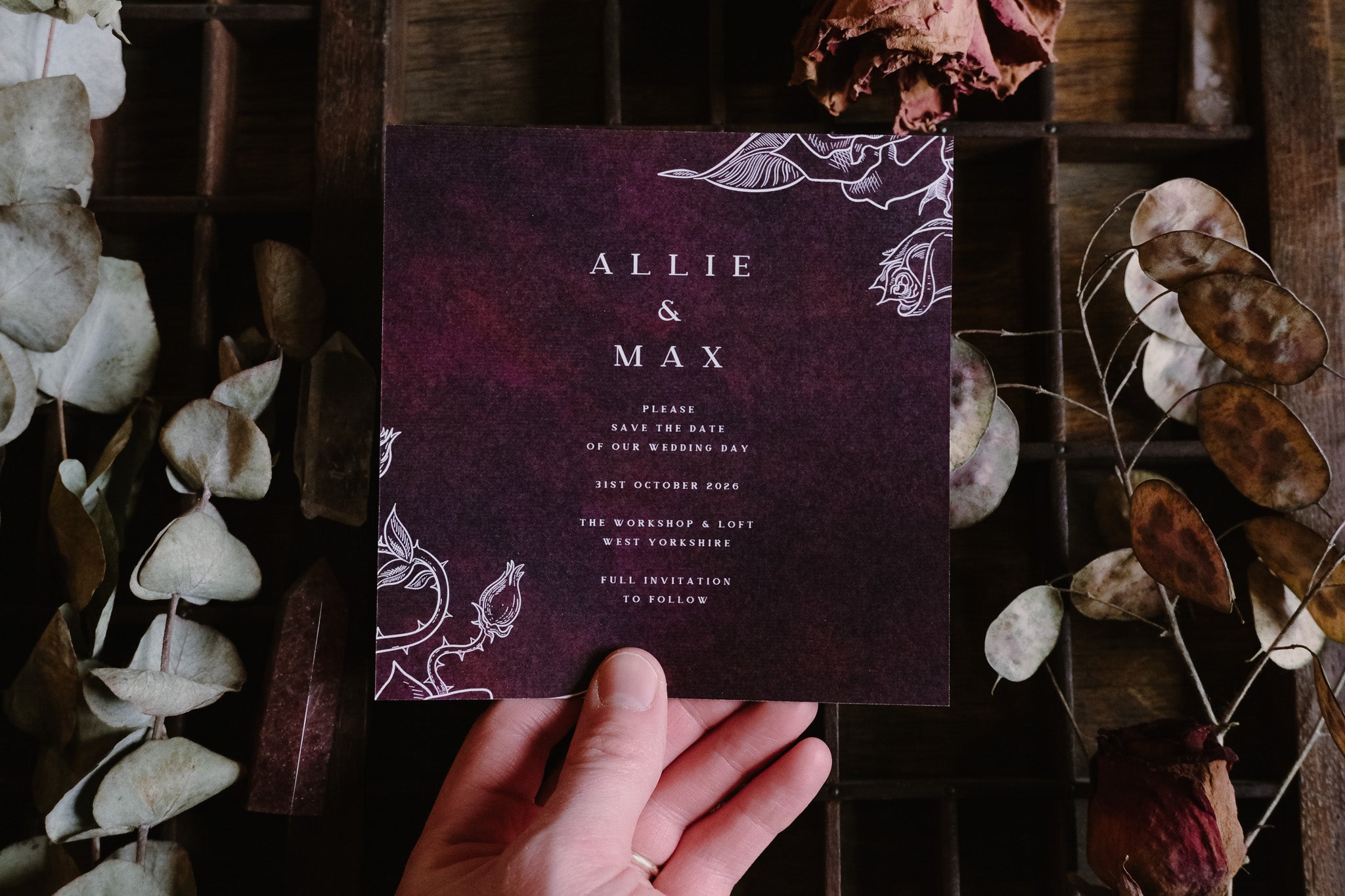 A hand holds a luxury gothic save the date card with dramatic dark floral design, surrounded by dried leaves; premium UK wedding stationery.