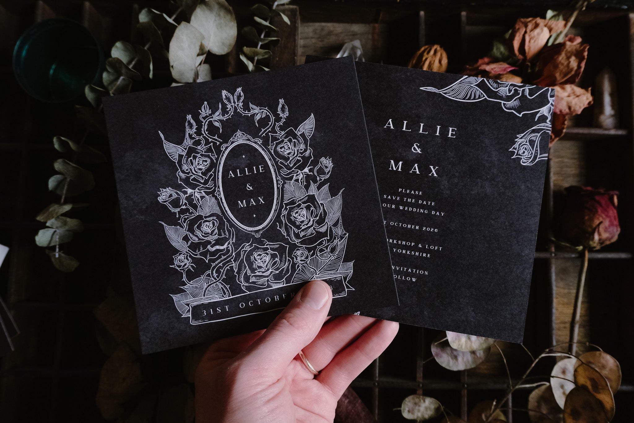A hand holds two black luxury wedding stationery cards with gothic white floral motifs, set amid dried boho flowers and leaves.