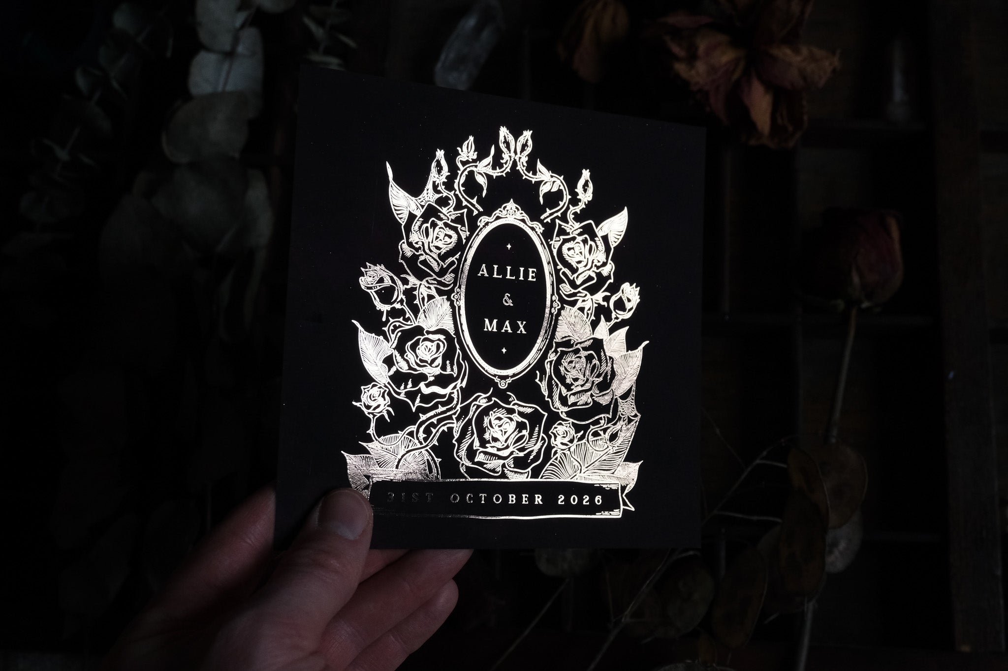 A hand presents a luxury black foiled gothic save the date card with floral accents, set against a dramatic dried flower backdrop.