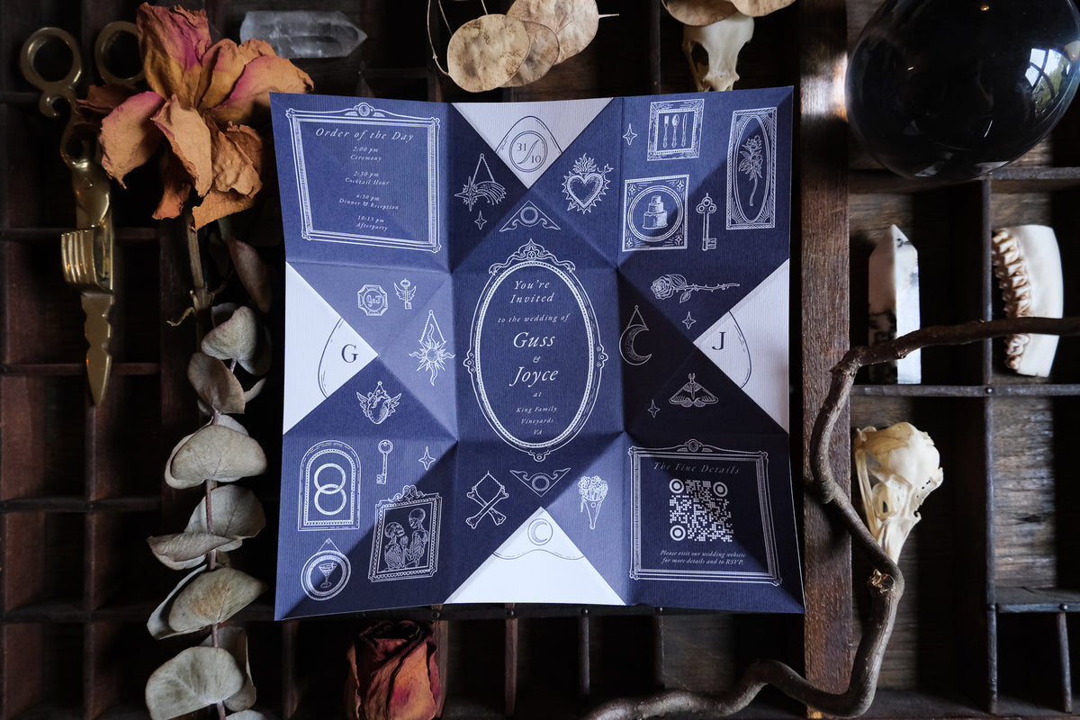 A Victorian-style folded wedding invitation with ornate white calligraphy, shown on a wooden tray with dried flowers and scissors.