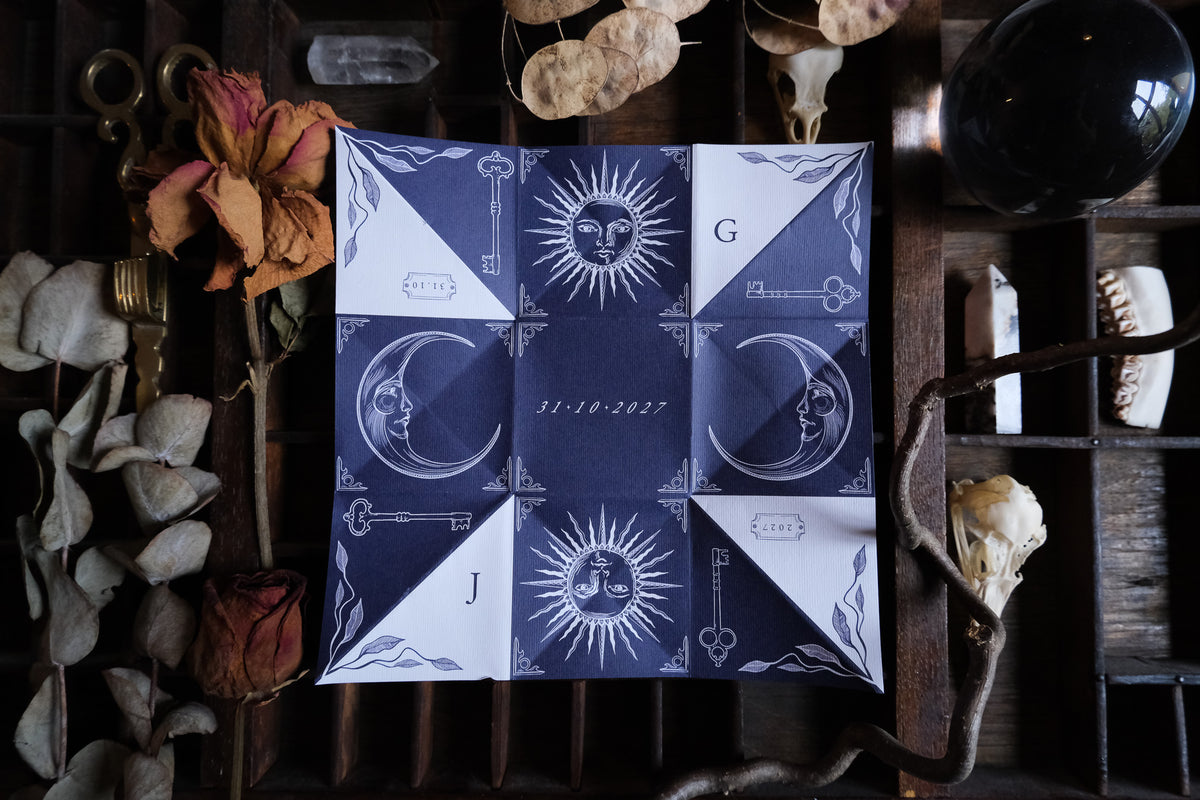 Victorian puzzle purse wedding invitation in soft neutral tones with celestial illustrations of suns, moons and keys, photographed folded on a lace tablecloth.