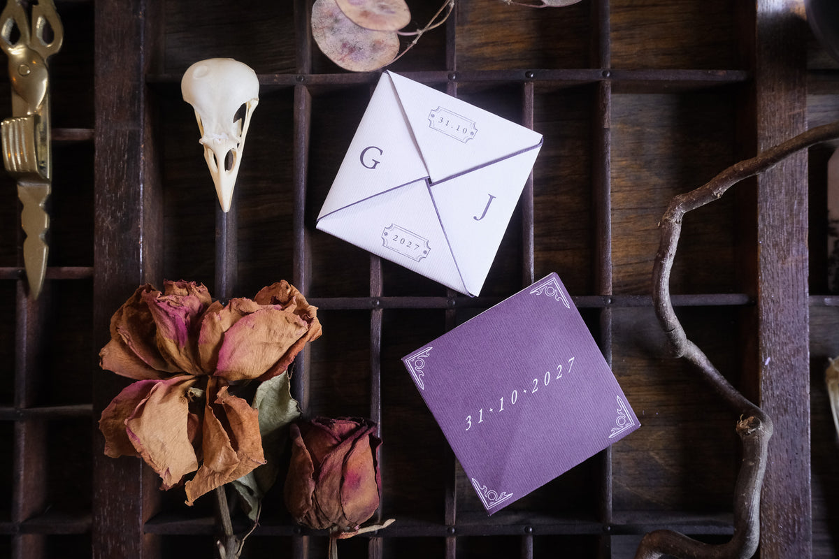 A Victorian style folding wedding invitation featuring illustrations of dried roses, an animal skull and a candle, arranged on a wooden grid.