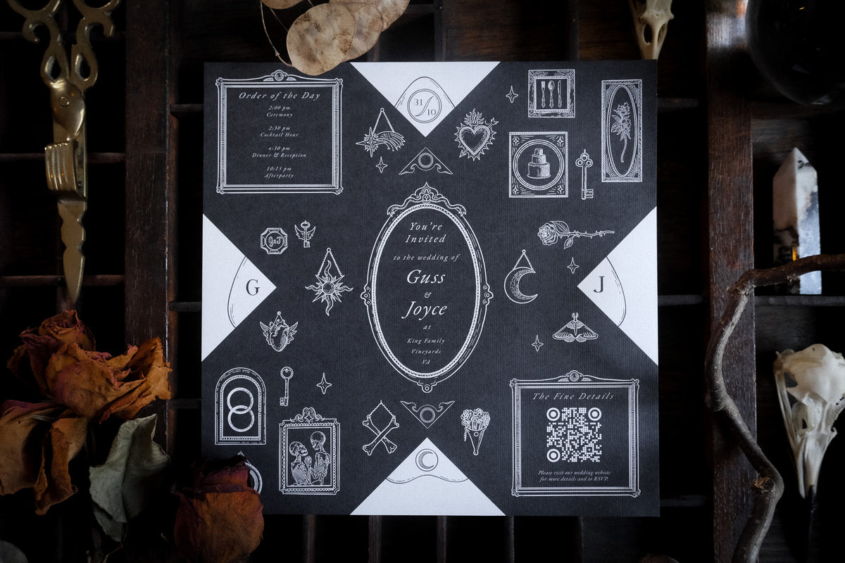 A Victorian style wedding invitation with white gothic illustrations and symbols on dark paper, photographed on a rustic table.