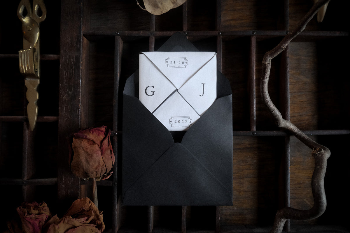 A vintage folded wedding invitation in a black envelope, displayed with dried roses and a wooden tray.