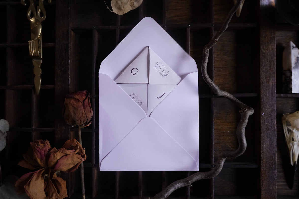 A Victorian puzzle purse wedding invitation in cream and muted tones, hand folded and displayed open on dark wood with dried flowers and twigs.