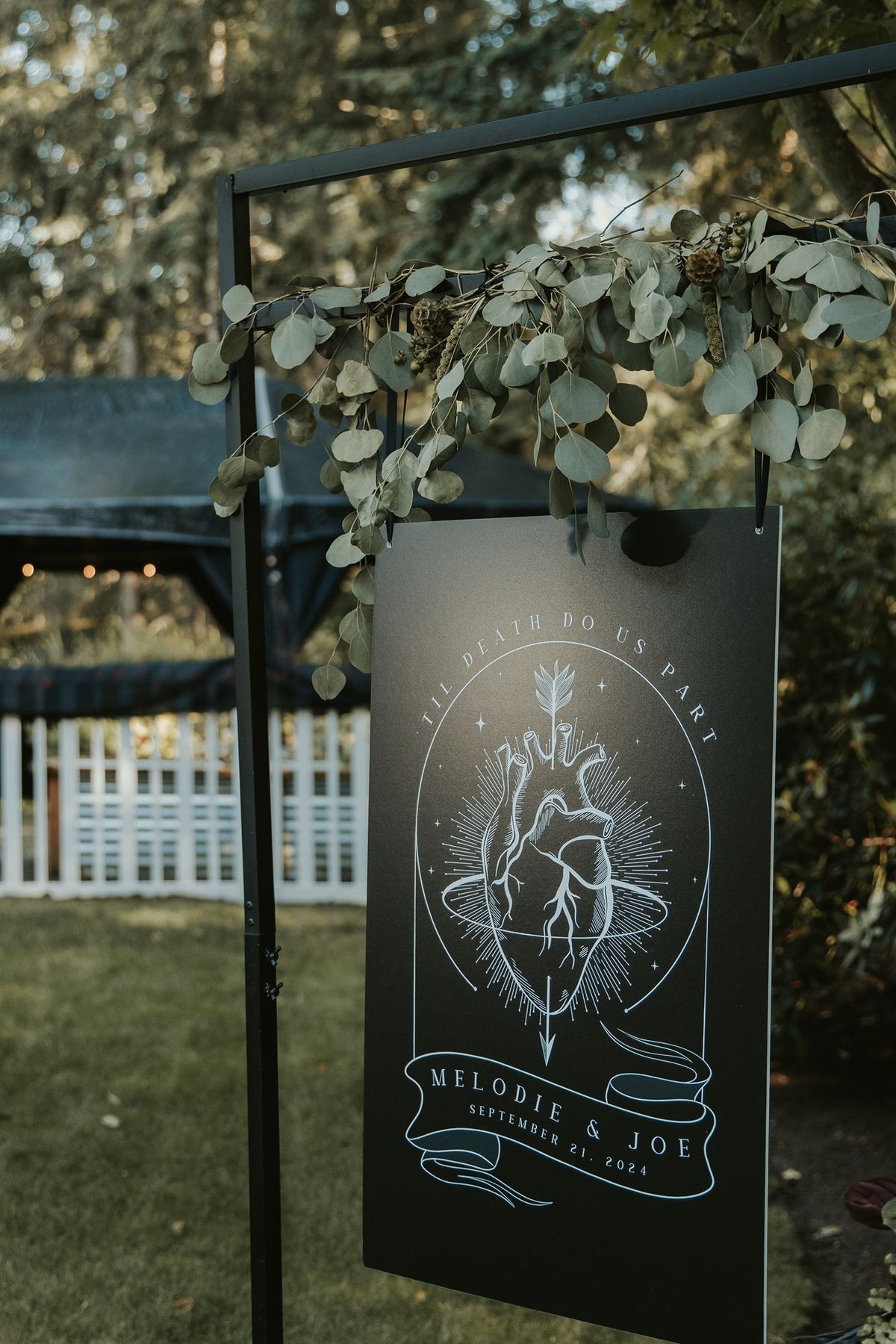 Gothic wedding welcome sign by Birdhouse197 with a red anatomical heart, rings, greenery and trees set in an outdoor scene.
