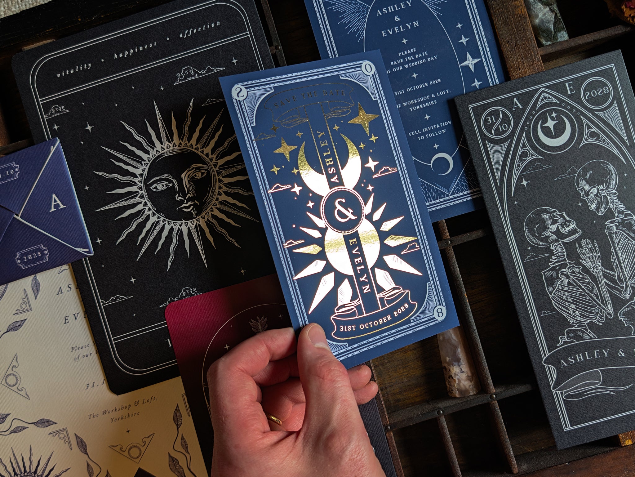 A hand holds an ornate blue luxury wedding stationery sample pack with celestial illustrations and gold foil detailing, evoking a mystical and romantic feel. Surrounding it are matching cards adorned with astrological and moon-inspired motifs, all arranged on a rustic wooden surface.