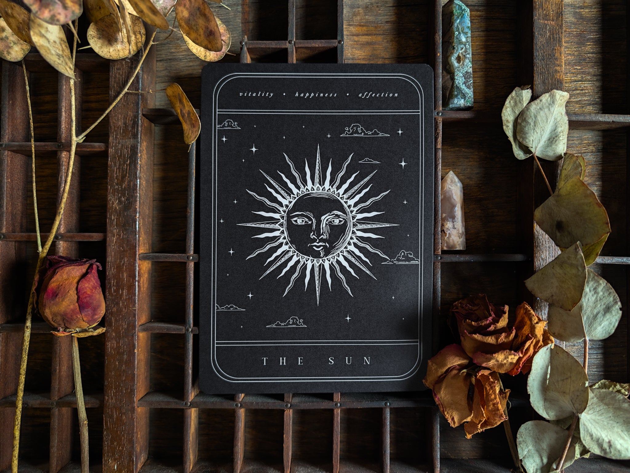 A luxury Save the Date wedding stationery sample pack is elegantly displayed, featuring a celestial-inspired card adorned with a radiant sun face illustration. The premium card is surrounded by dried flowers and leaves, set against a rustic wooden grid background for a romantic, boho atmosphere.