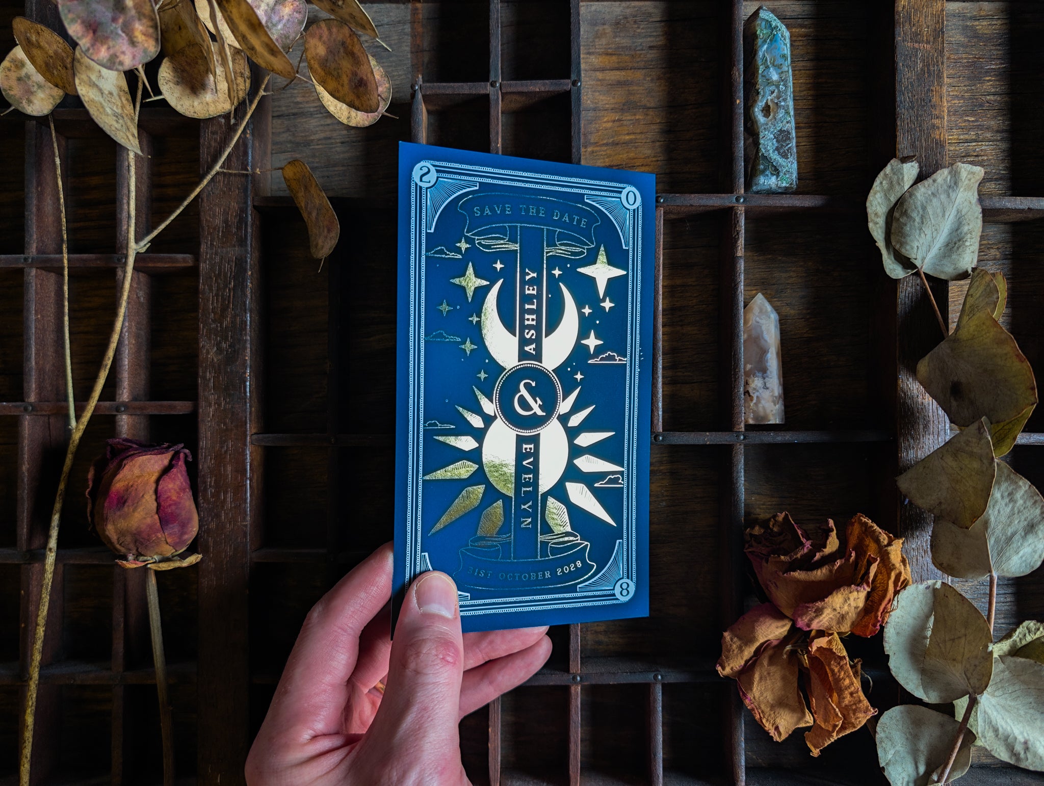 Dusk & Dawn celestial save the date card in gold foil on blue, held in hand.