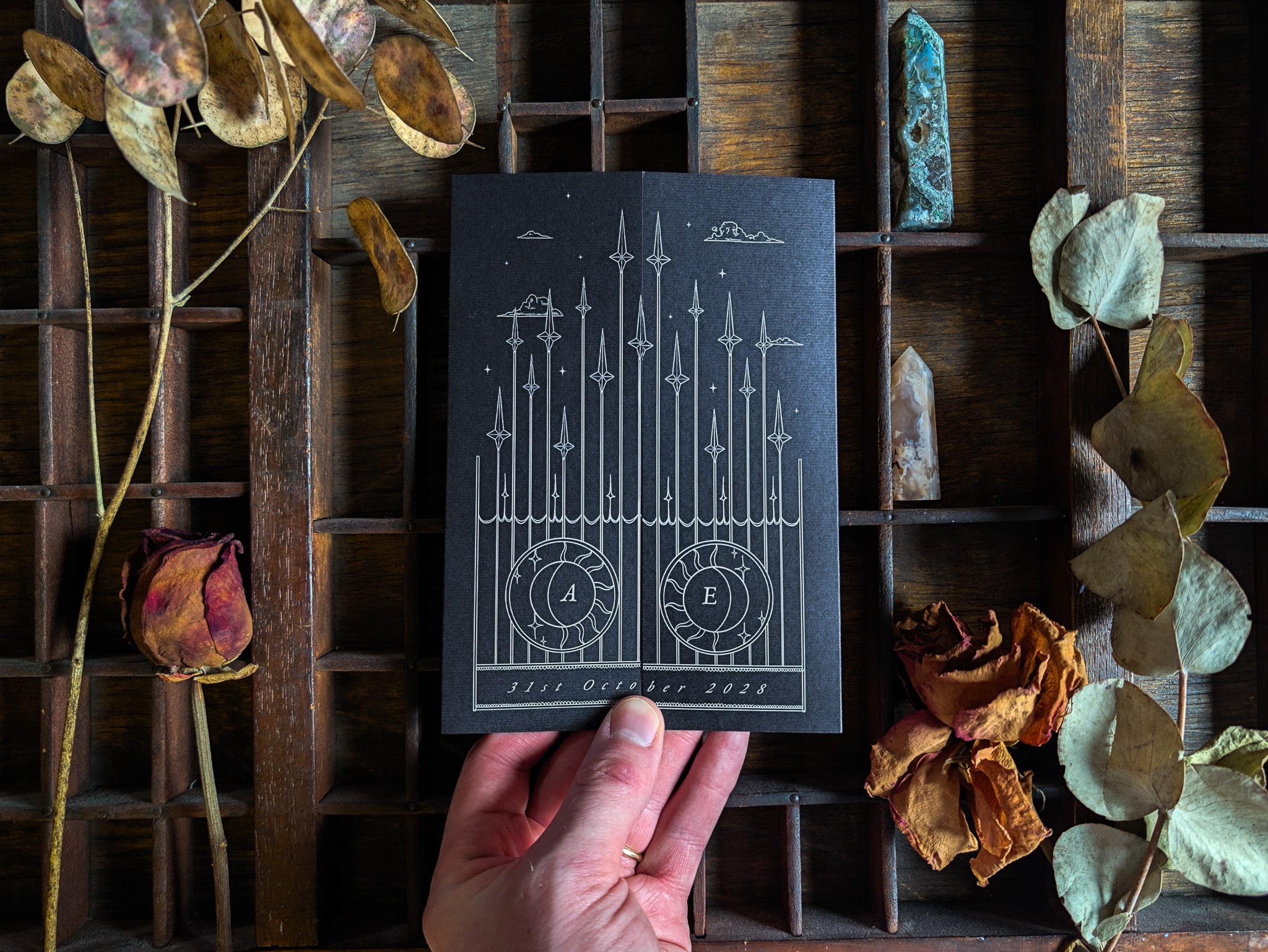 The Skeleton Lovers gatefold invitation in matte black card, held in hand.