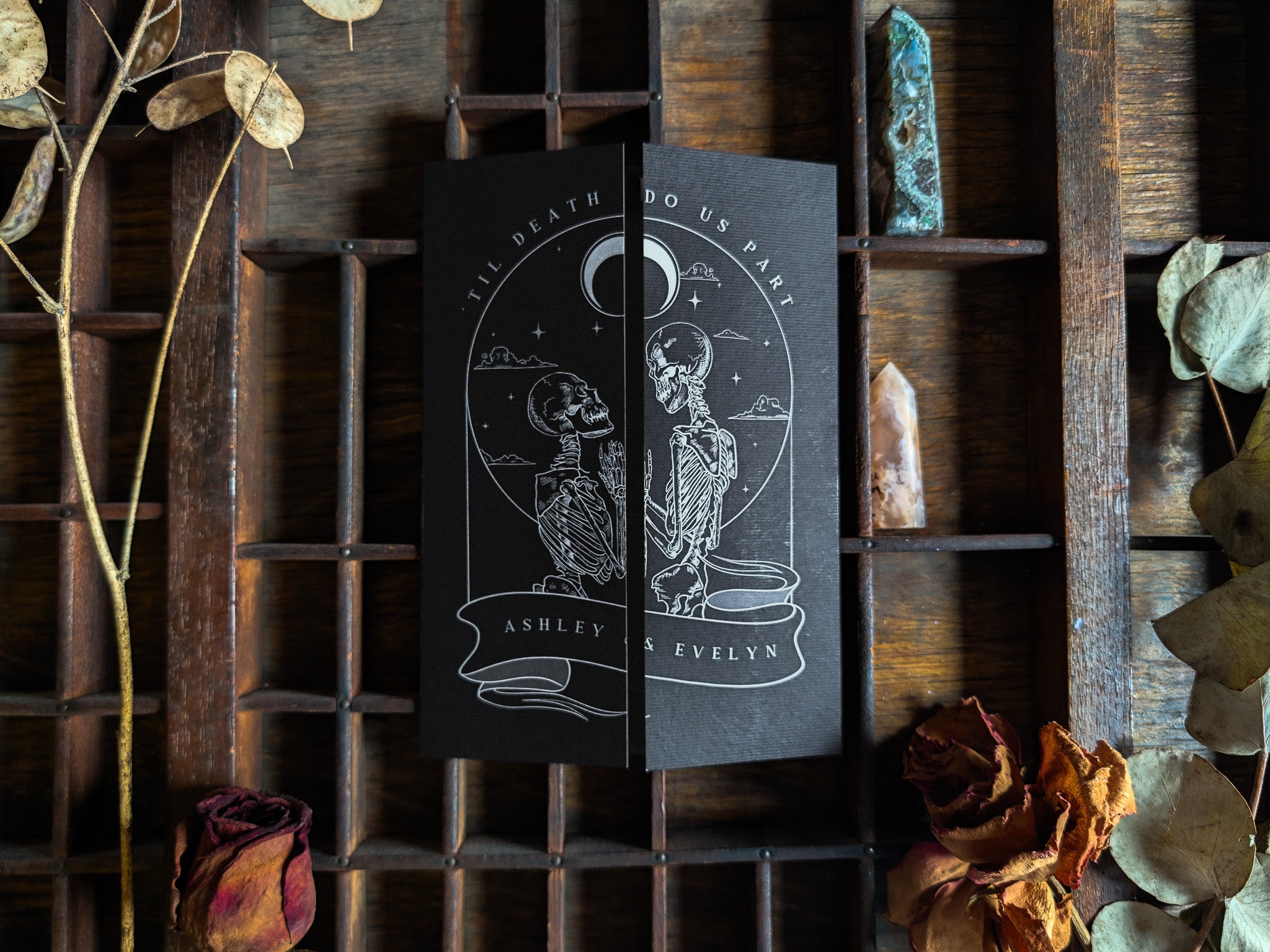 Premium UK wedding stationery sample pack featuring a dramatic black invitation with a gothic, alternative design. The card displays two intricately illustrated skeletons facing each other beneath a celestial moon motif, creating an aura of romantic mystique. Surrounded by dried flowers and crystals in a carefully styled setting, this luxury wedding invitation set evokes vintage and boho influences ideal for couples seeking unforgettable, visually bold stationery inspiration.