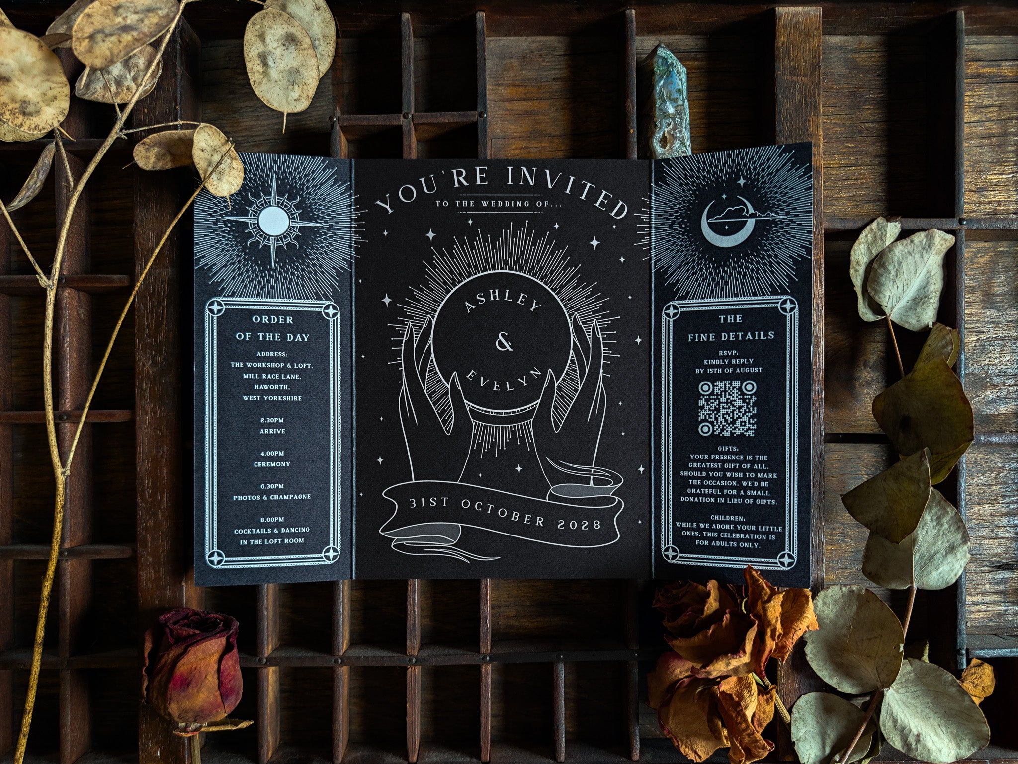 A luxurious black wedding invitation in a celestial and hand-illustrated design is elegantly presented on a rustic wooden tray, accented by dried flowers and leaves. This dramatic gatefold sample from a premium UK stationery collection exudes alternative gothic style and vintage romance, perfect for couples seeking mystical wedding stationery with artistic flair.