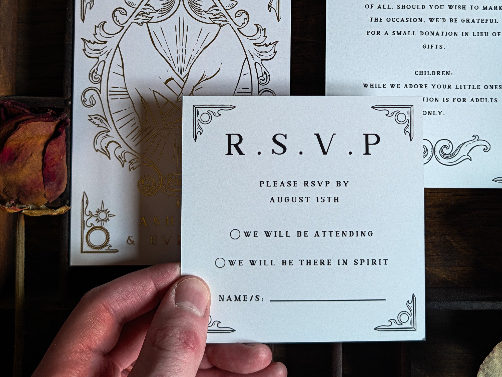 A hand displays a luxury RSVP card from a premium UK wedding stationery suite, crafted in a romantic style with delicate foil accents and personalised details. In the background, matching foiled invitations are artfully arranged alongside a dried rose, creating an elegant and timeless setting.
