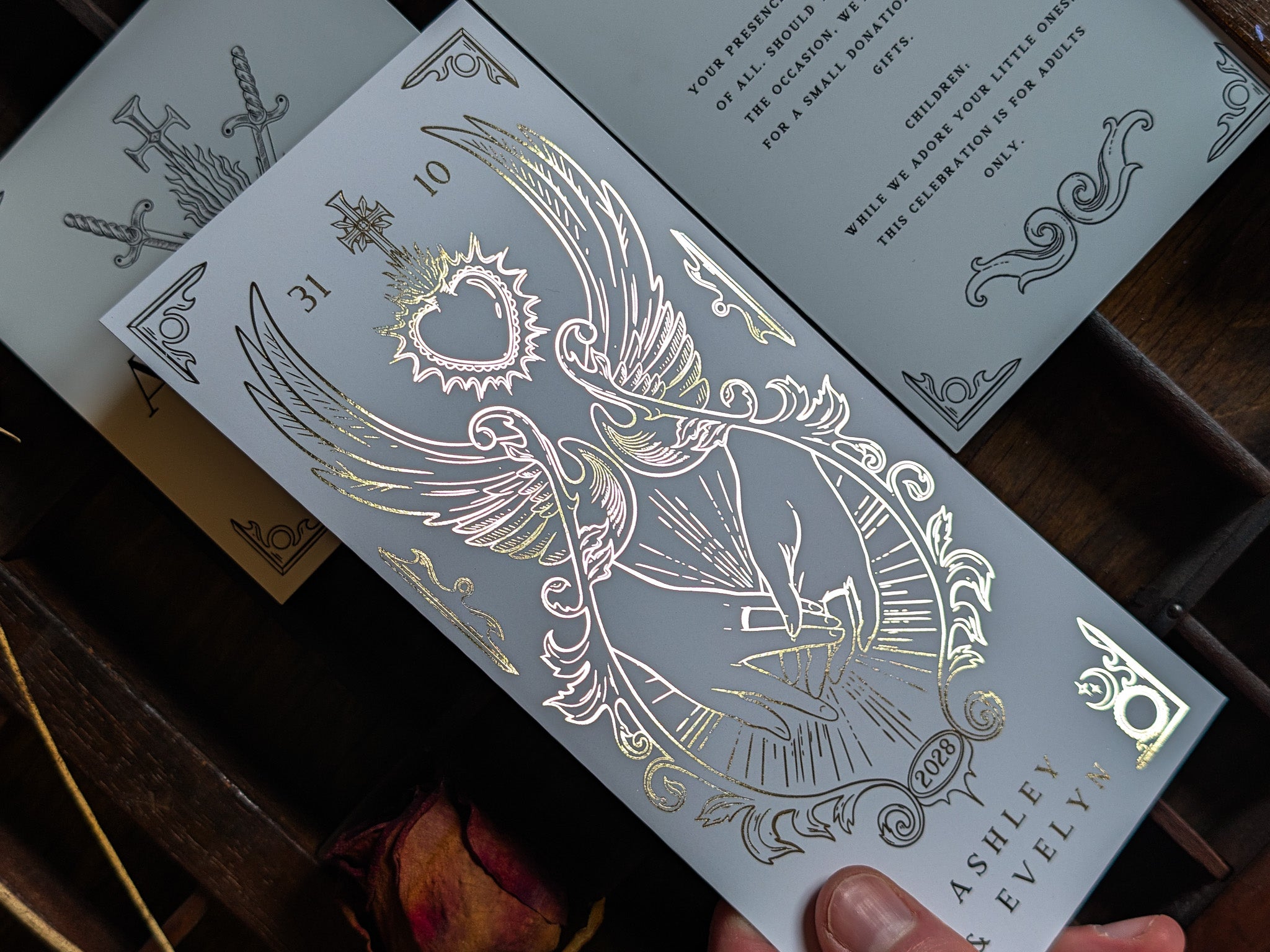 A luxury silver foil-embossed wedding invitation featuring an intricate winged heart motif, exuding a dramatic and gothic elegance, is displayed on a rustic wooden surface and partially overlaps an ornate grey gatefold design.