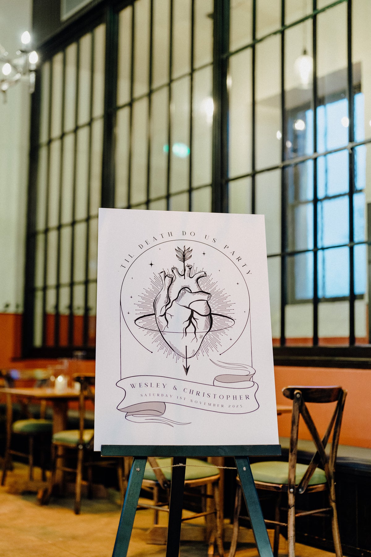 A gothic wedding welcome sign by Birdhouse197 with a red anatomical heart illustration, displayed on an easel in a cafe setting.