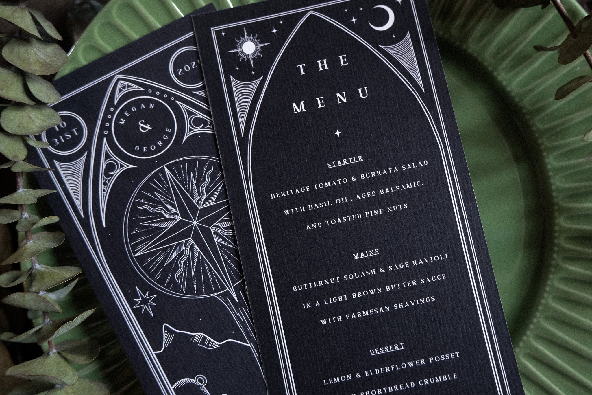 Luxury UK wedding menu card with a dramatic celestial tarot-inspired black design, white illustrations, on fern-accented plate.