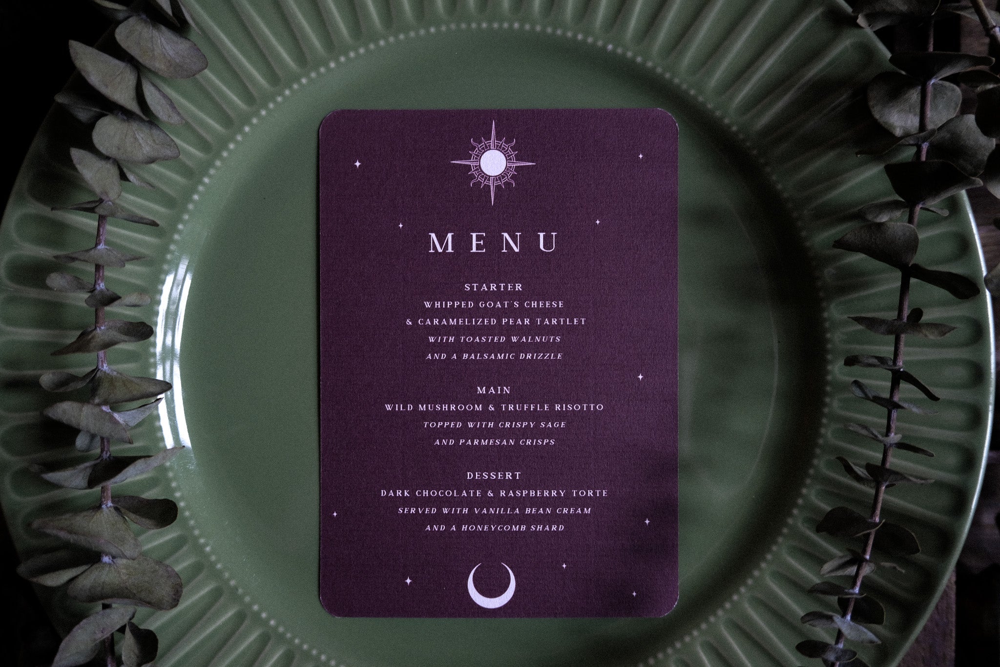 Luxury wedding menu cards with a gothic, romantic design and celestial accents, styled on green plates with eucalyptus leaves.
