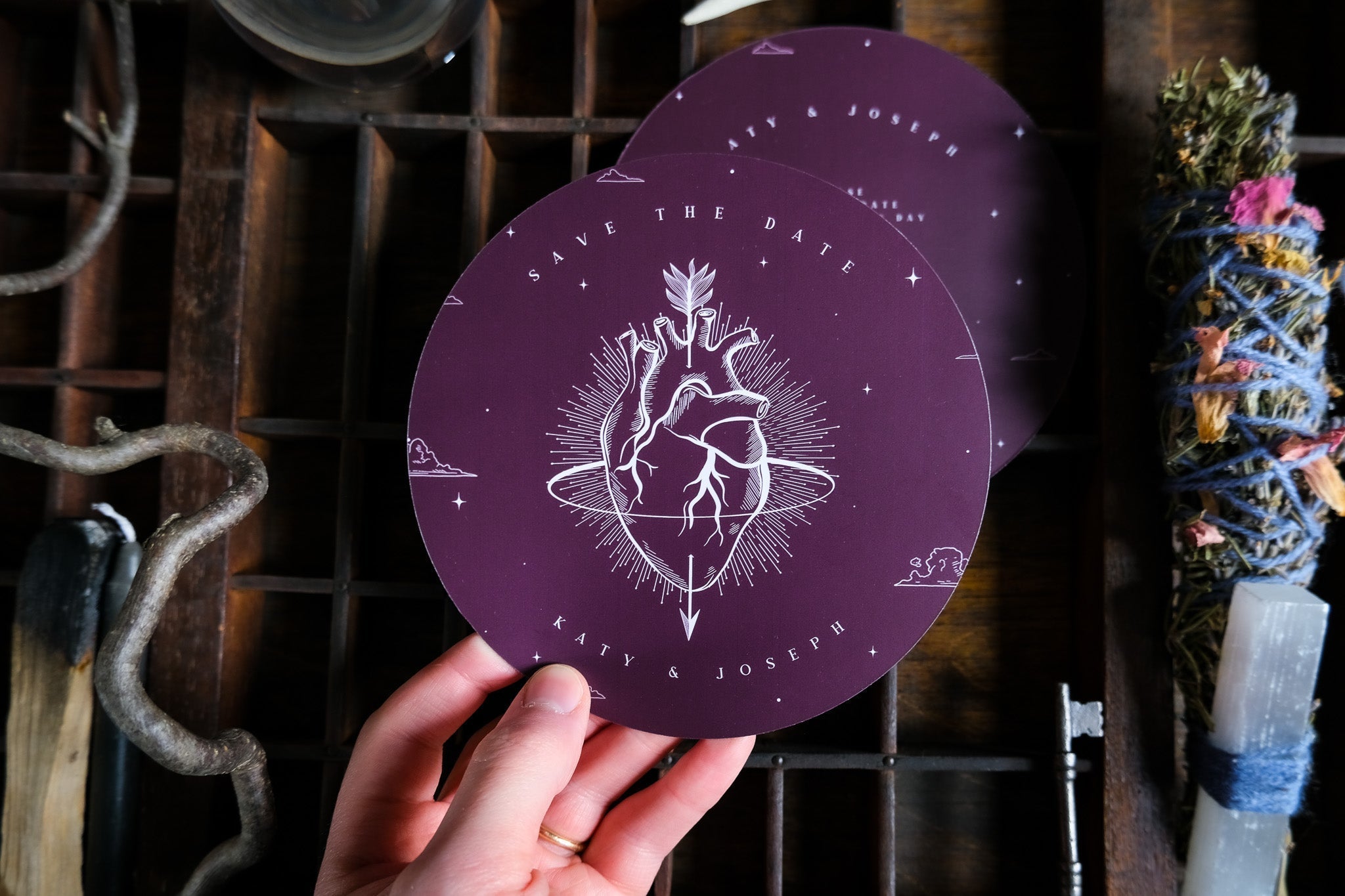 A hand holds a luxury gothic circular save the date card with an anatomical heart, arrow, and celestial stars; dramatic boho stationery and mystical decor lie nearby.