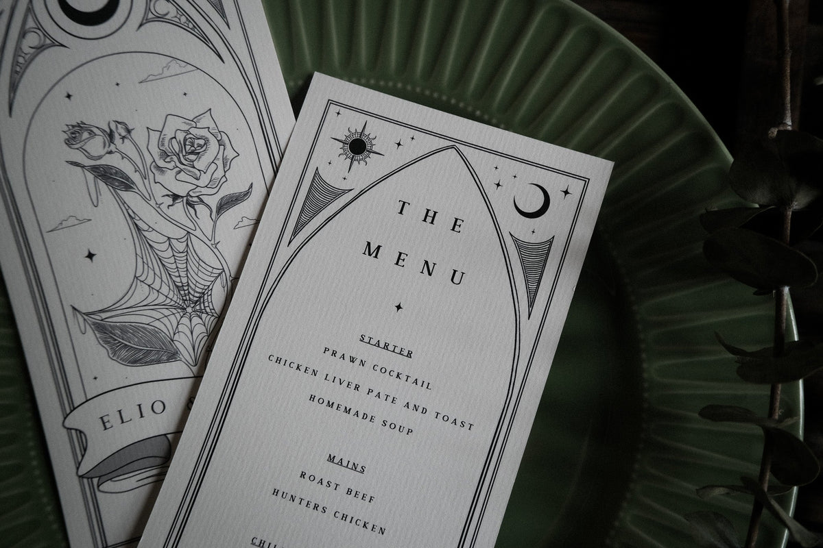 Close-up of luxury UK wedding menu cards with gothic romantic design, monochrome rose and spiderweb art, elegant type, dramatic appeal.