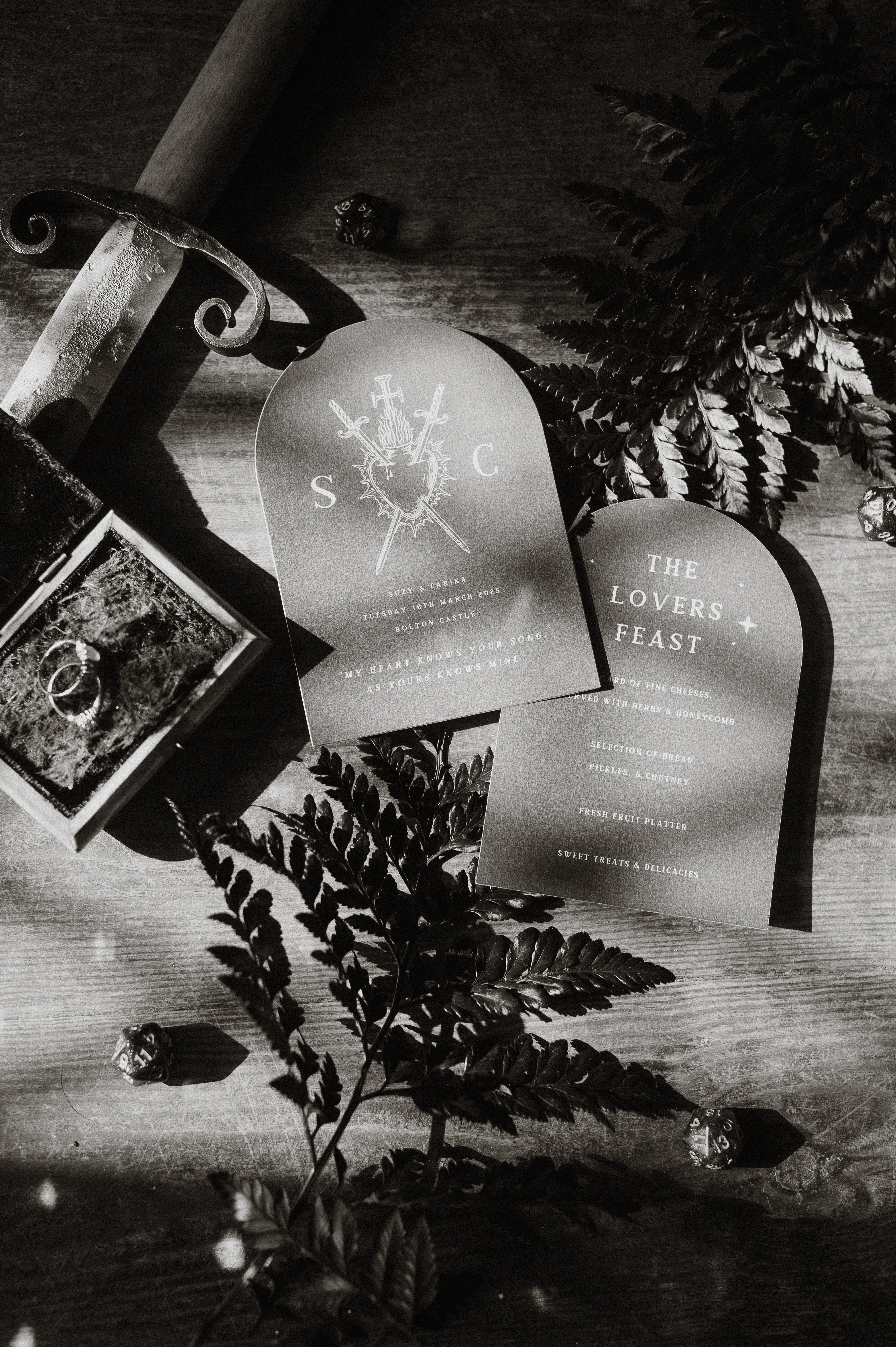 Black and white image of two gothic-inspired wedding invitations on wood, styled with ferns, rings, sword hilt, and dice.