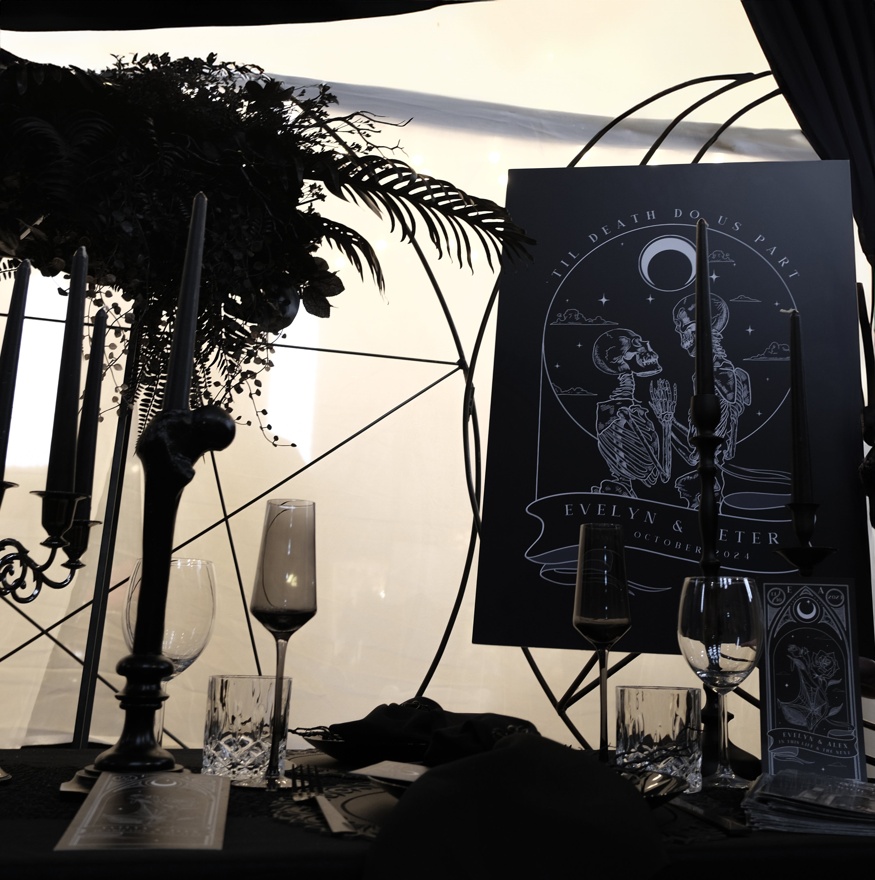 A luxury gothic wedding welcome board with tarot-inspired skeleton couple art, set on a dramatic table with black candles and florals.